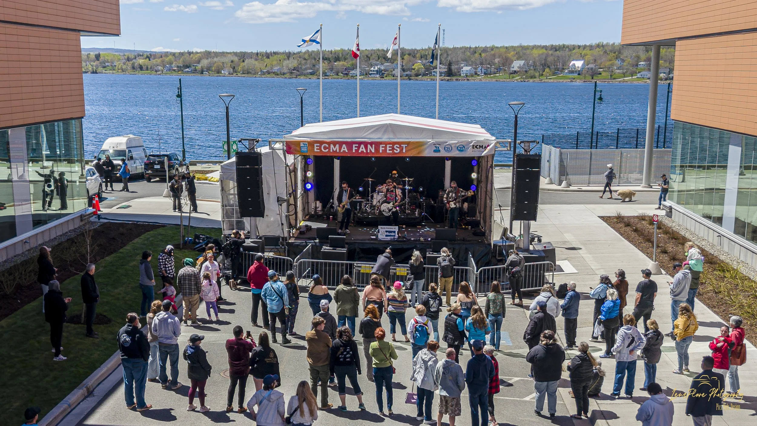 An outdoor concert takes place by a large body of water with a band playing on a stage labeled "ECMA FAN FEST". People are watching the performance from the ground, some standing and some walking. Flags are flying above the stage, and some cars are p