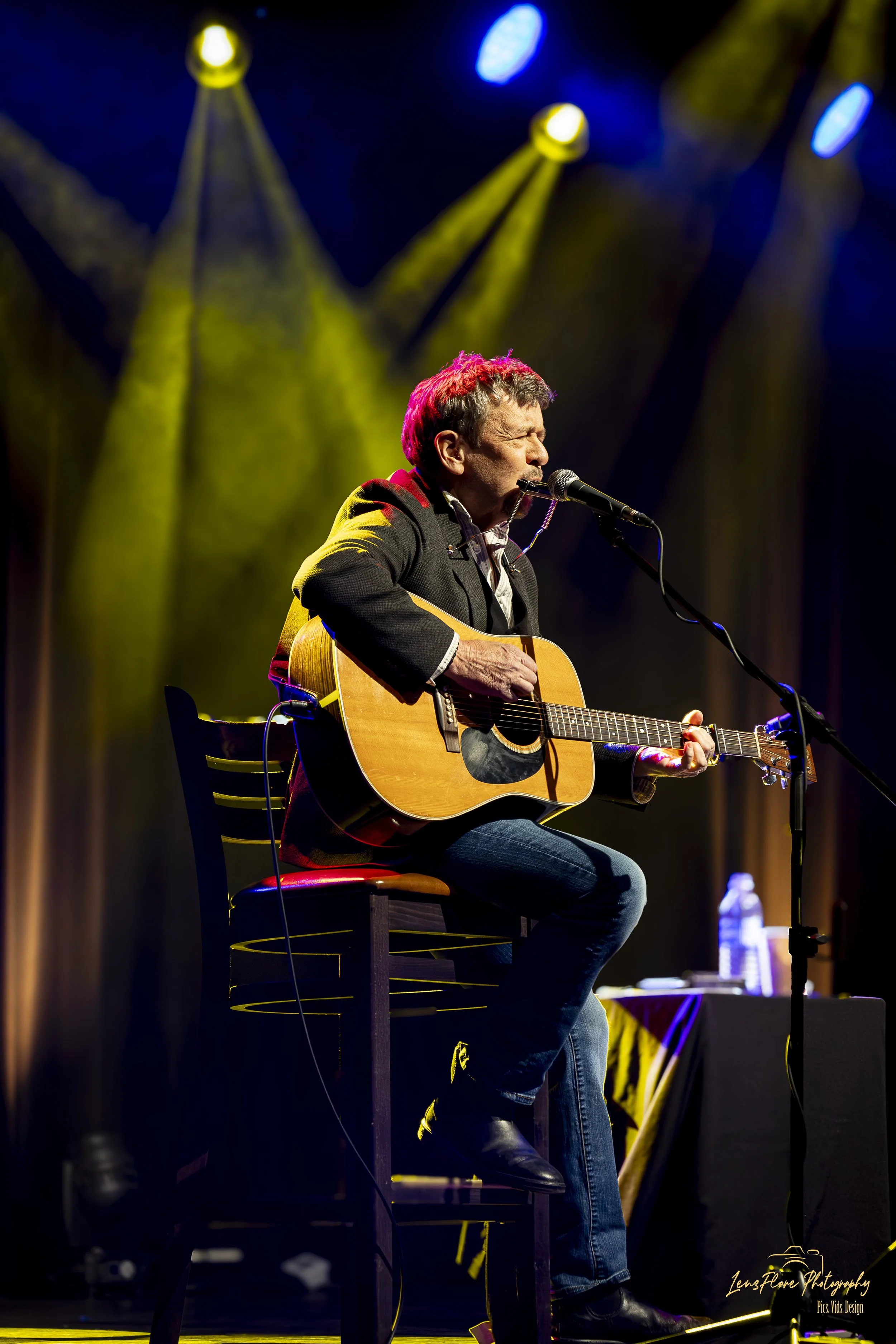 A man with gray hair wearing a black jacket and jeans, sitting on a wooden stool, singing into a microphone and playing an acoustic guitar on stage. The stage has yellow and blue spotlights focused on him, with a dark background.