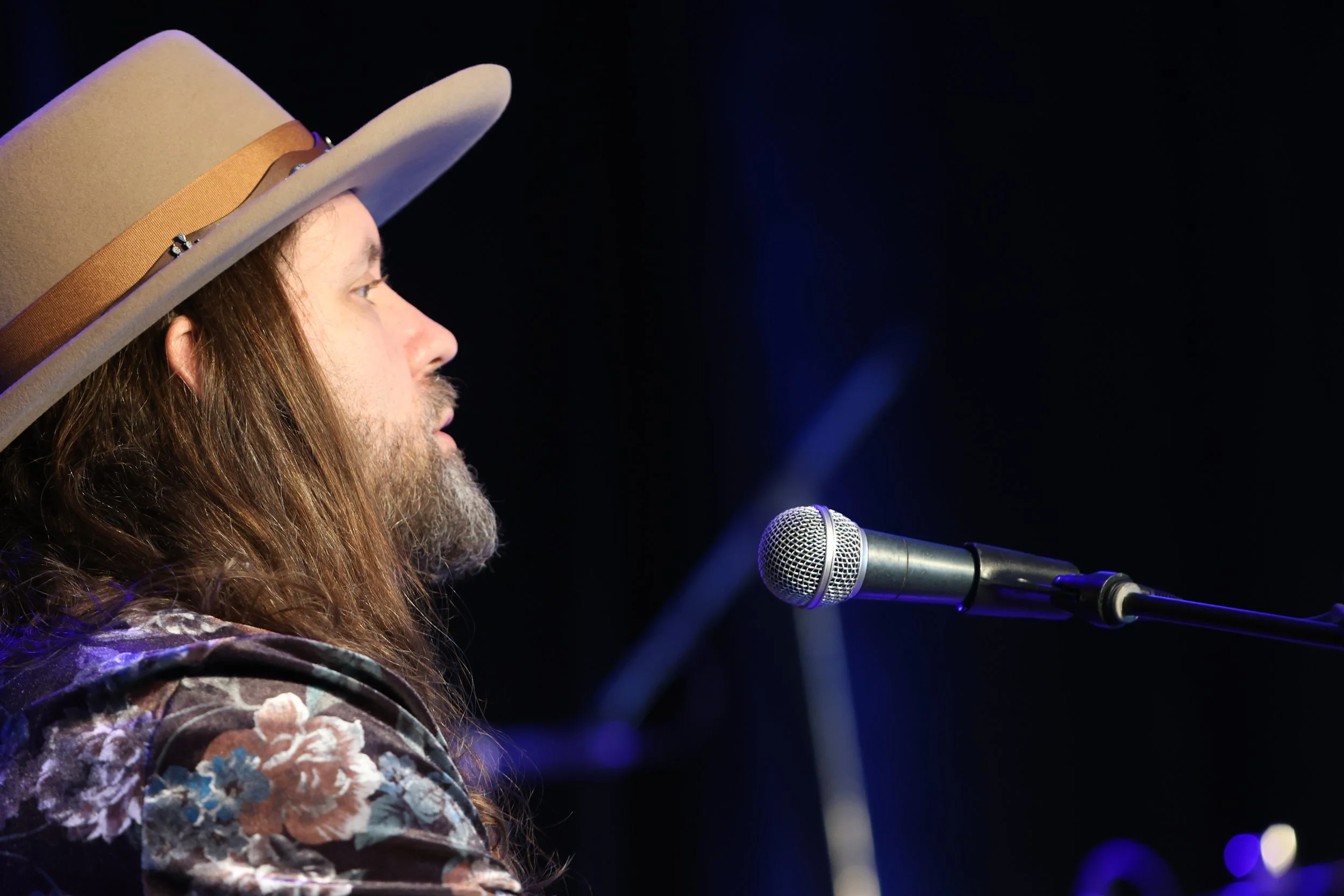 Side view of a man with long hair and a beard singing into a microphone on stage, wearing a beige hat and floral shirt.