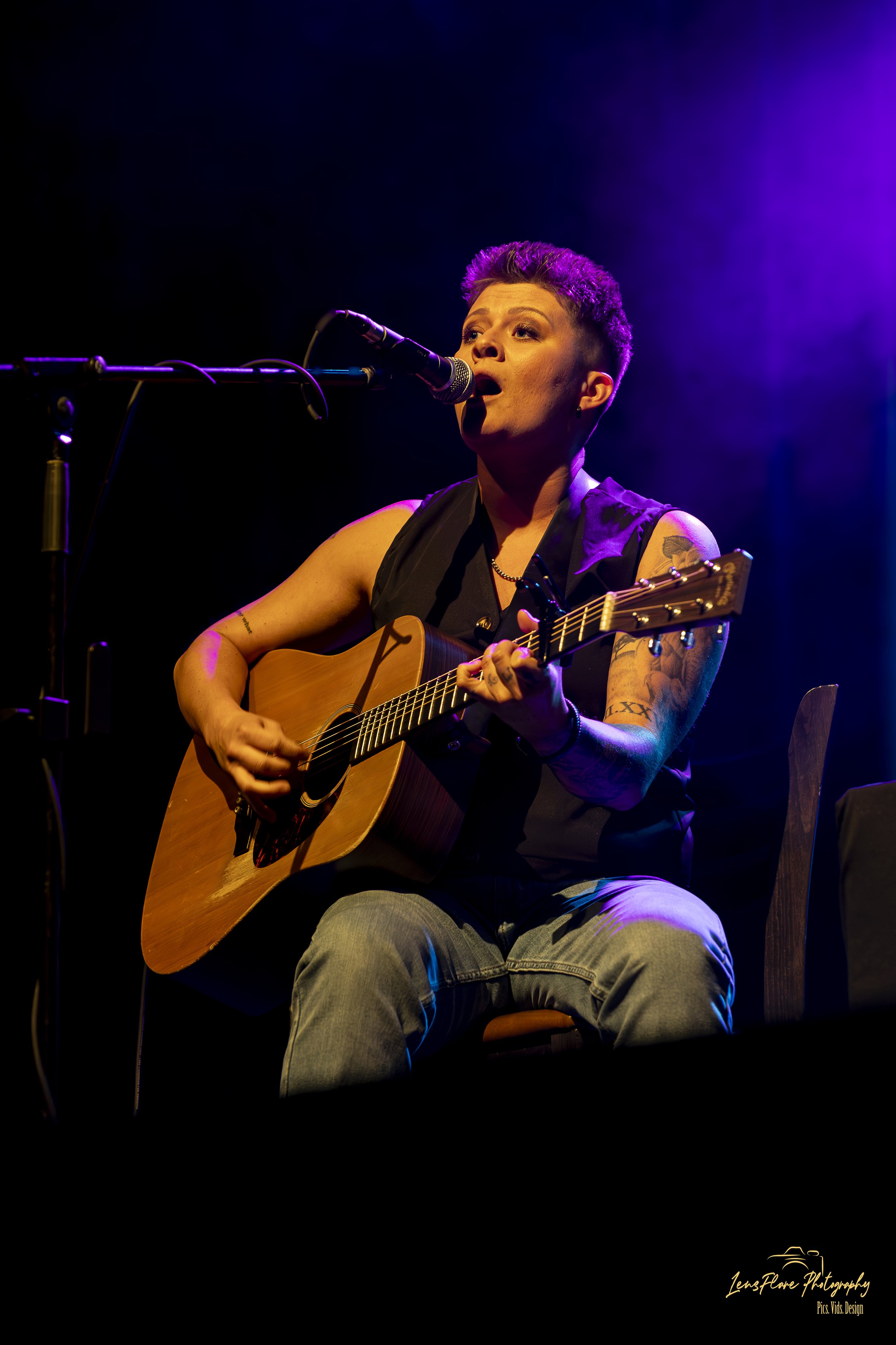 A woman with short, purple dyed hair singing into a microphone while playing an acoustic guitar on stage, illuminated with purple and yellow stage lighting.