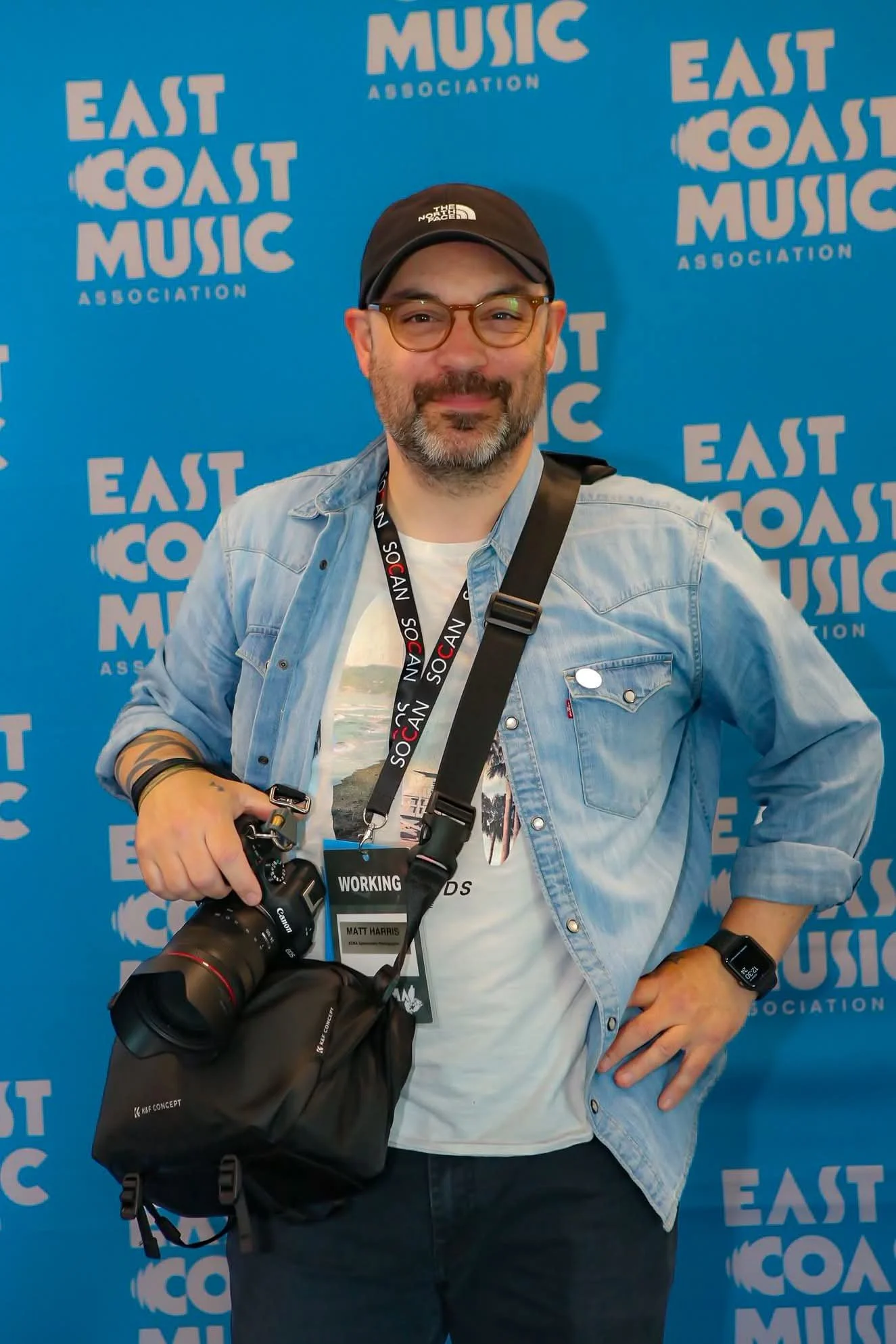 A man with glasses and a beard smiling, standing in front of a blue background with 'East Coast Music Association' logos, holding a camera with a strap around his neck, wearing a denim jacket, a black cap, and a wristwatch.