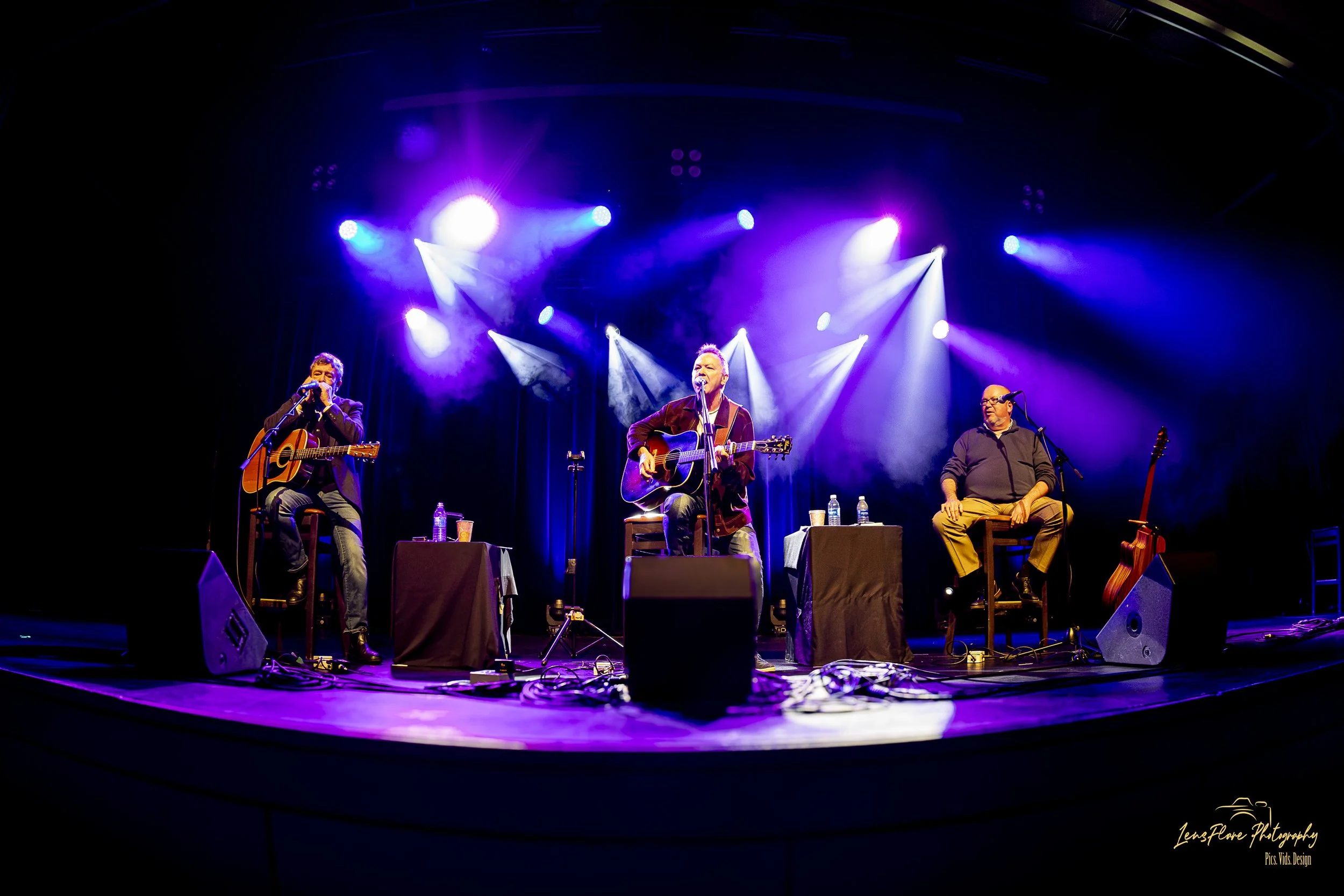 Three musicians performing on stage with purple and white stage lighting. Two guitarists are seated with guitars, and one man with glasses and a microphone is sitting on a stool. There are water bottles and a music stand on stage.