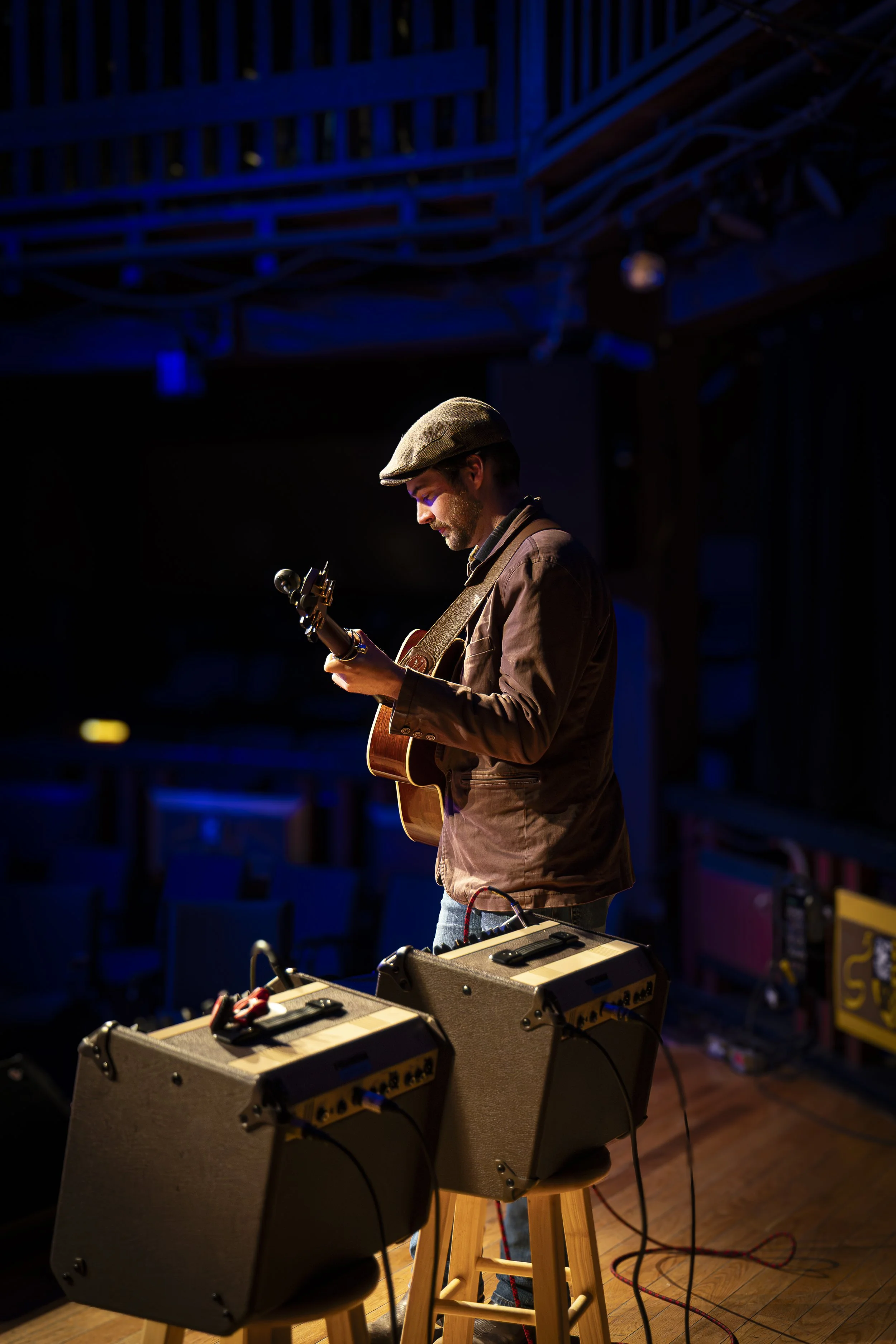 A musician wearing a flat cap and brown jacket playing an acoustic guitar on stage, with equipment and cables in front of him and blue stage lighting overhead.