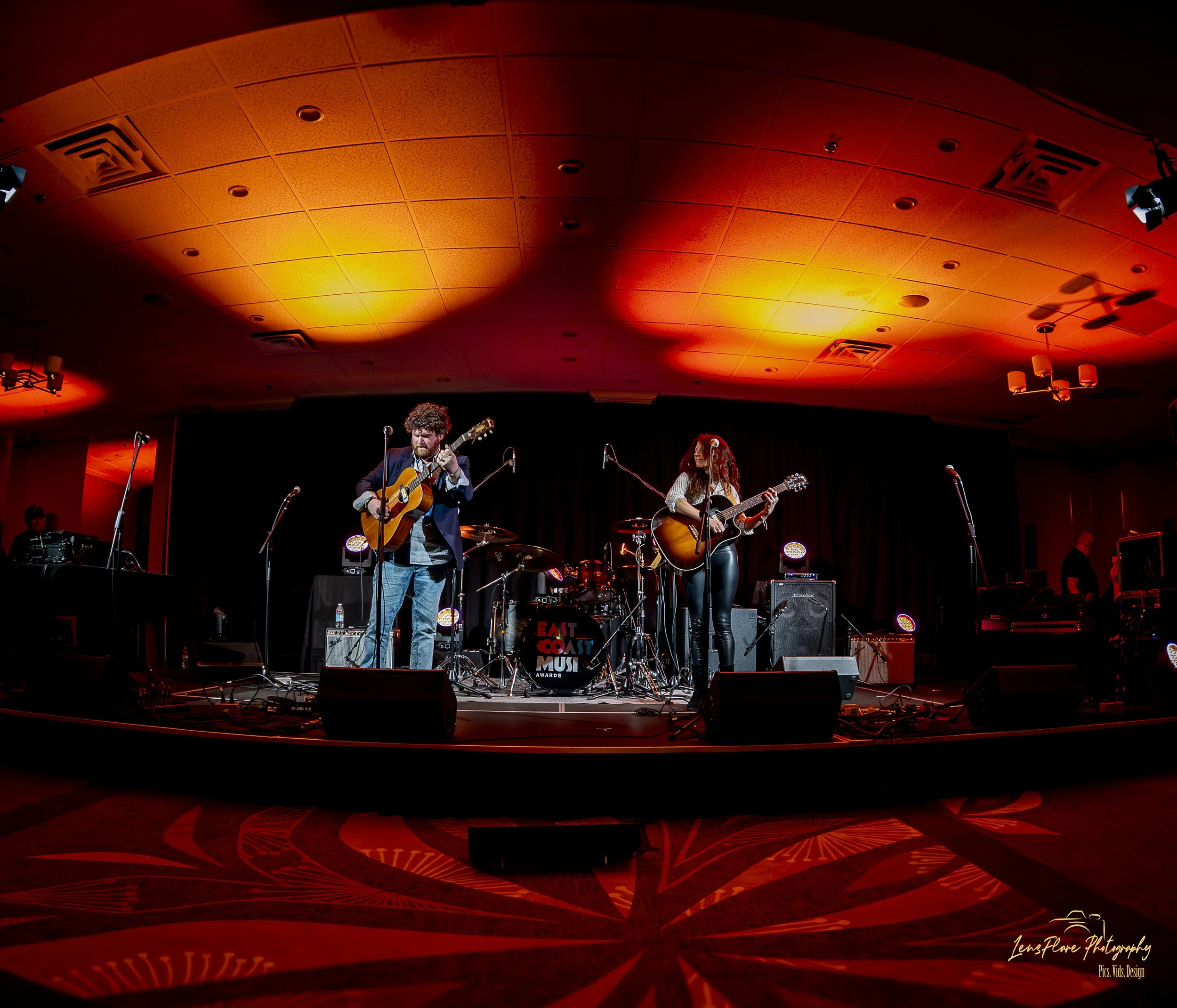 Musicians performing on stage under orange and red lighting, with one man playing guitar and singing, and a woman also with a guitar singing, at the East Coast Music Awards event.