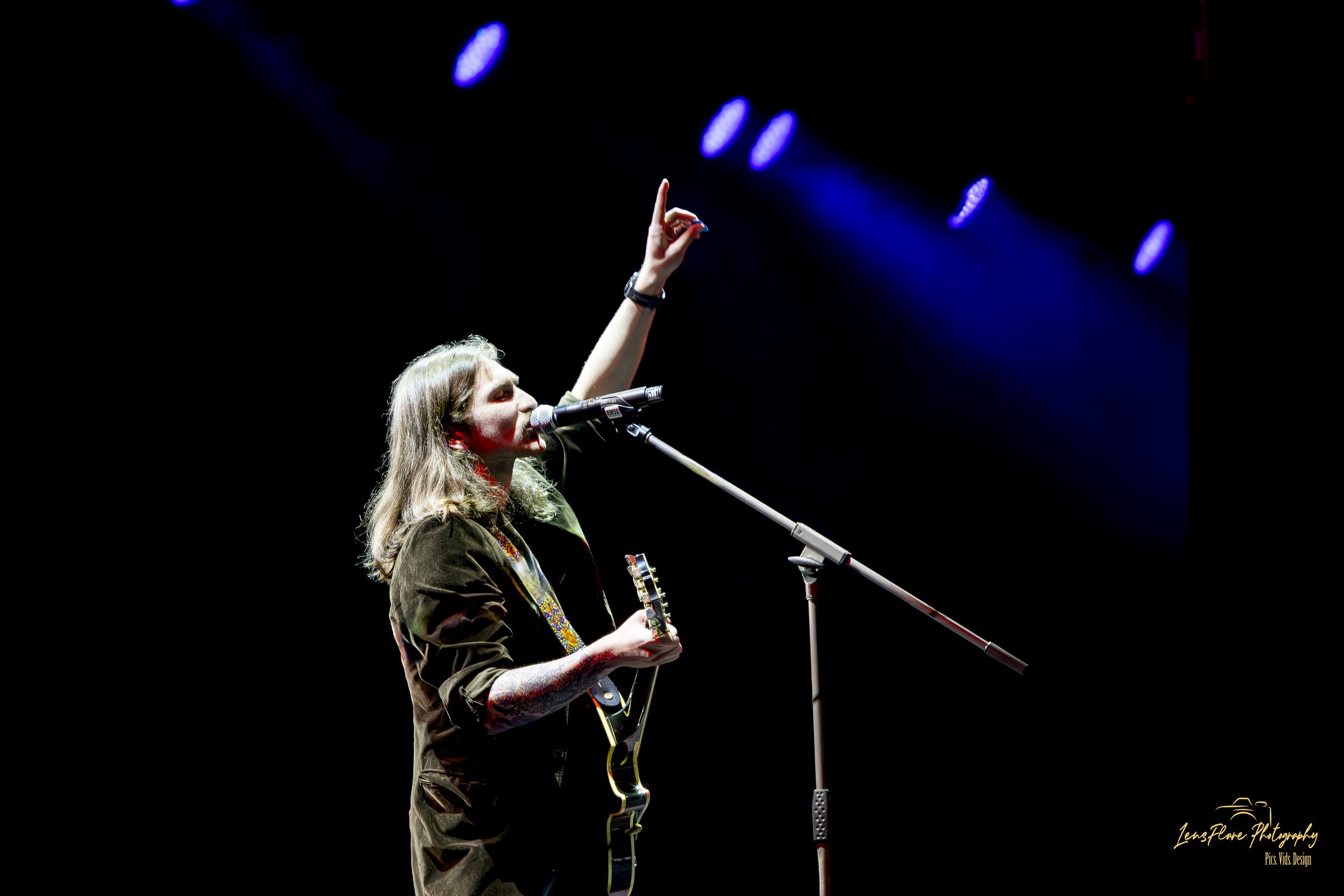 A woman with long hair singing into a microphone while playing an acoustic guitar on stage, raising her hand with one finger pointing upward. The background is dark with blue stage lights.
