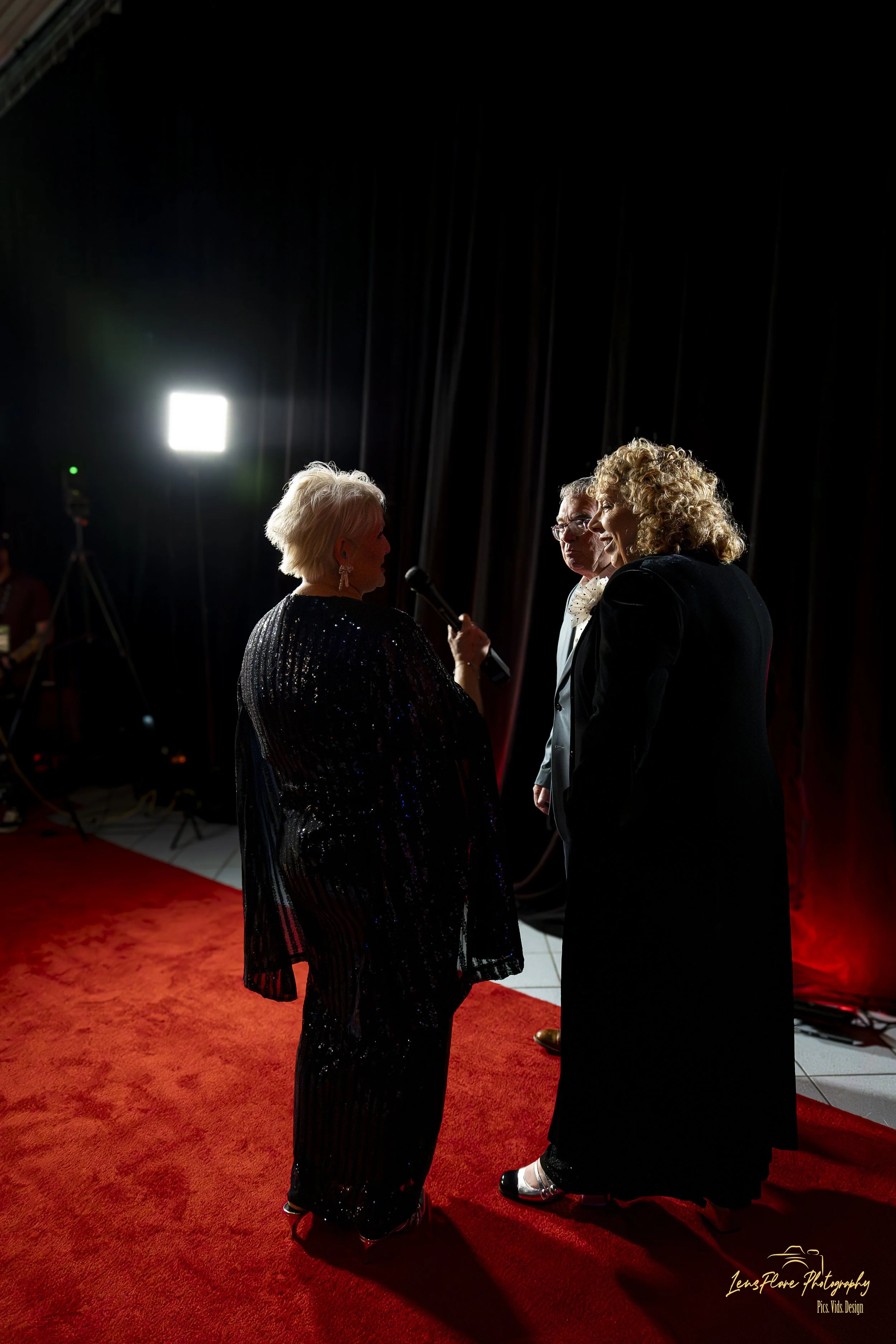 Three people on a red carpet at an event, with two women and one man, engaged in conversation in front of black curtains and bright lighting.