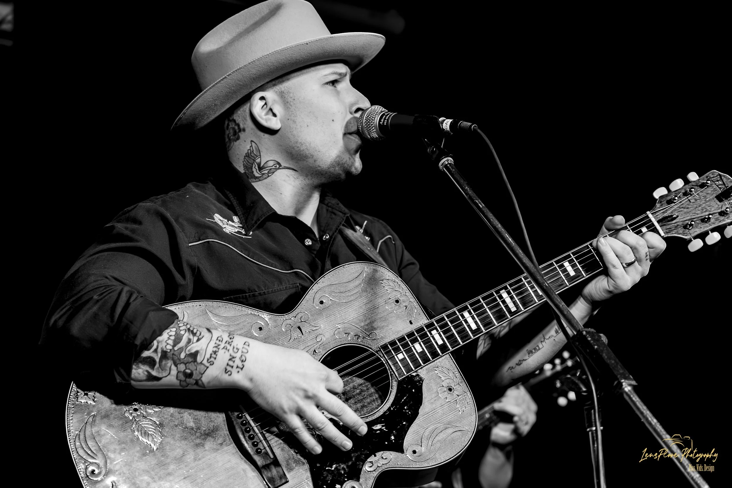 A black-and-white photo of a man singing into a microphone while playing an acoustic guitar. He is wearing a hat and has tattoos on his arm and neck.