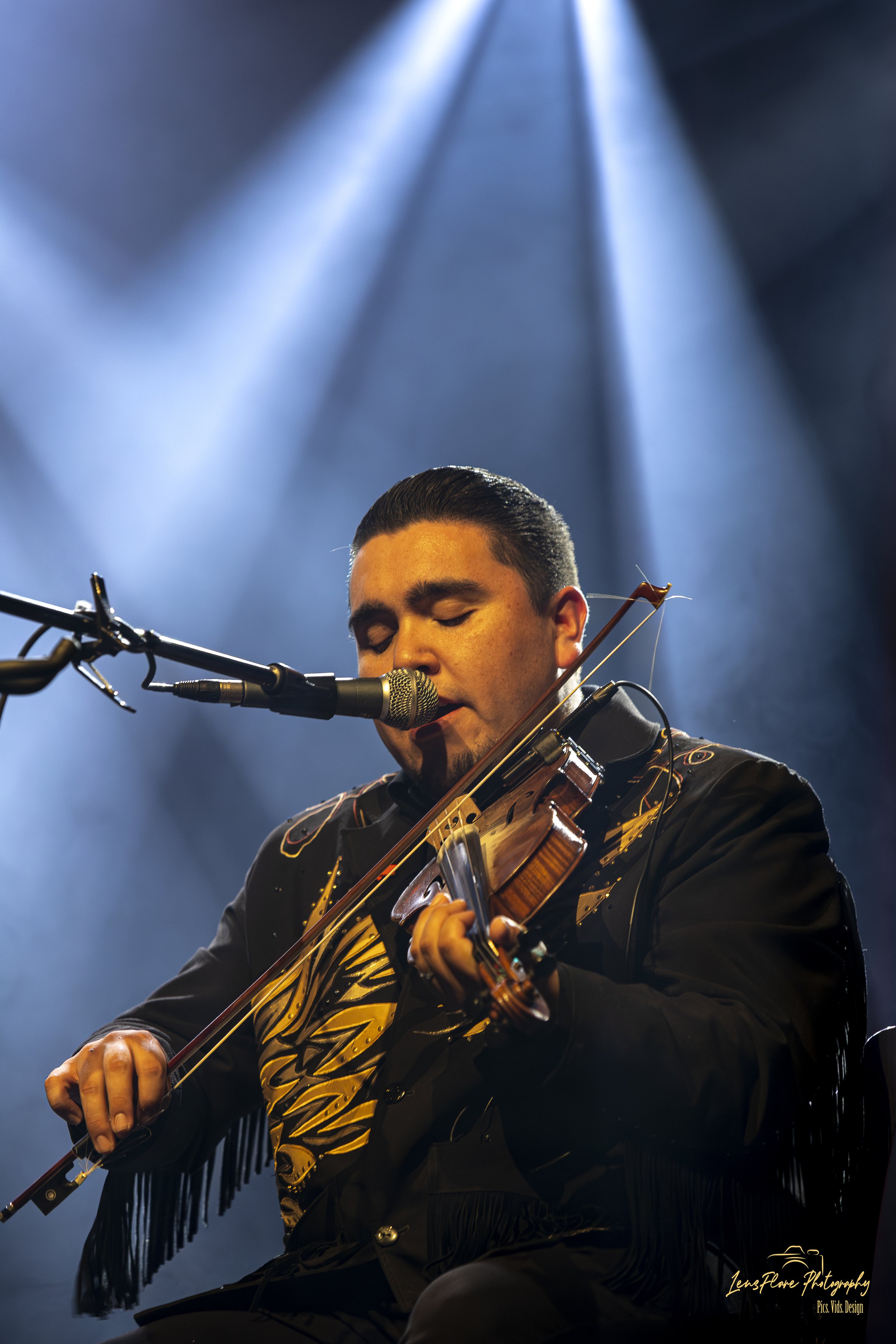 A man playing the violin on stage, illuminated by stage lights, with his eyes closed and dressed in a black jacket with fringe details.
