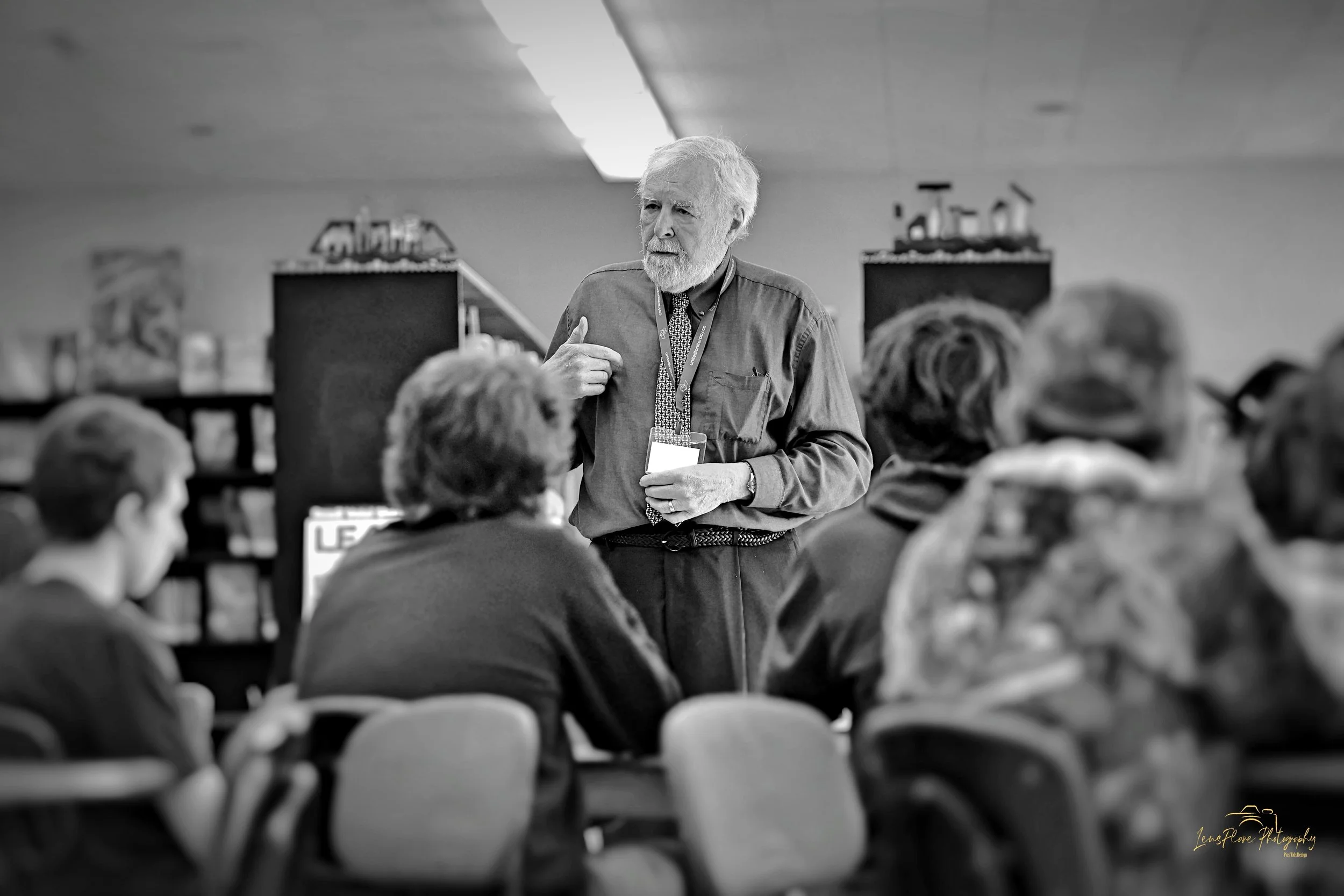 An older man with white hair and beard giving a presentation or lecture to a group of seated women in a room that appears to be a library or community center, with shelves and books in the background.