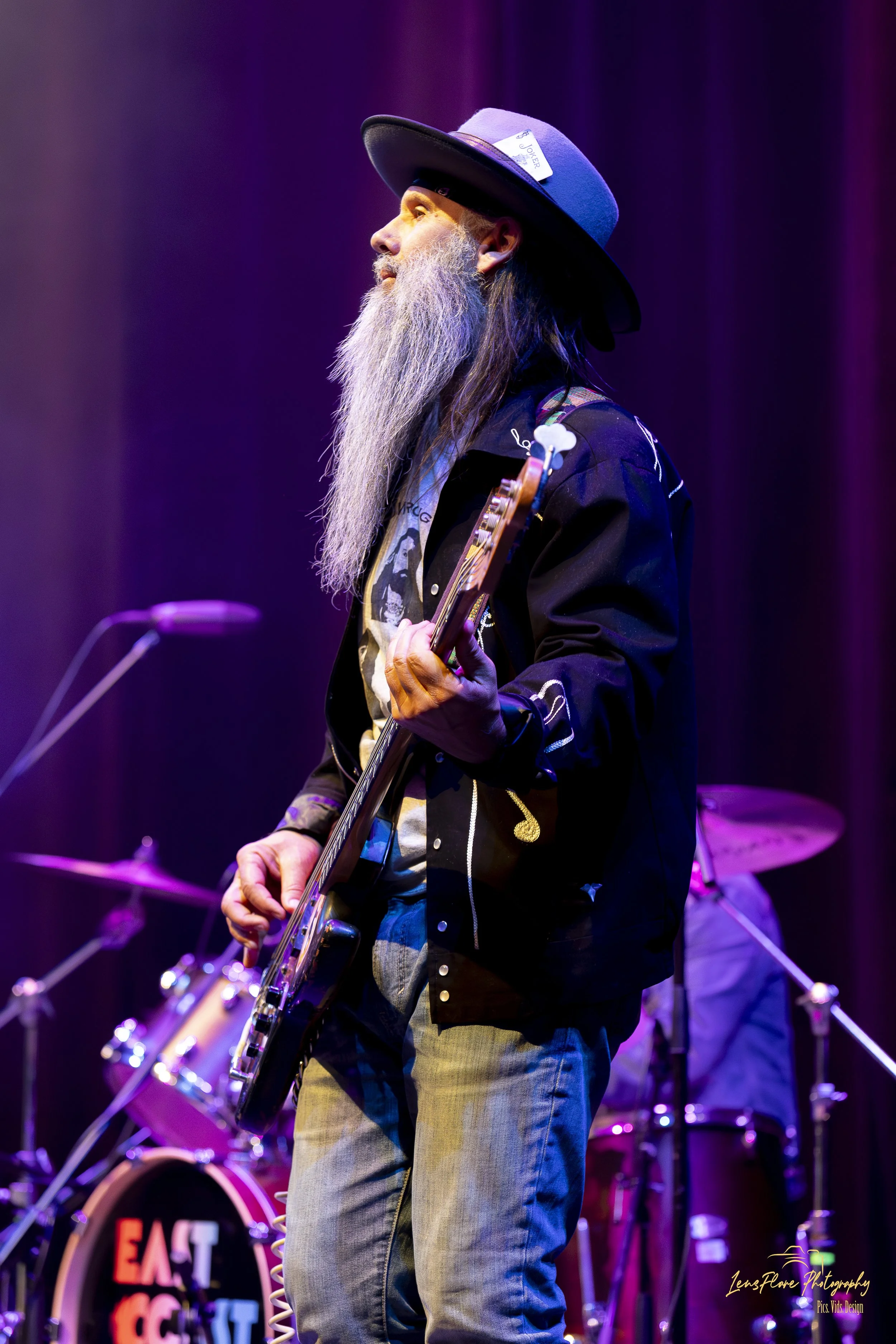 A man with a long gray beard and long hair, wearing a black hat with a Joker card tucked into it, playing an electric guitar on stage surrounded by purple and blue lighting. There are drums and a sign with partially visible text in the background.