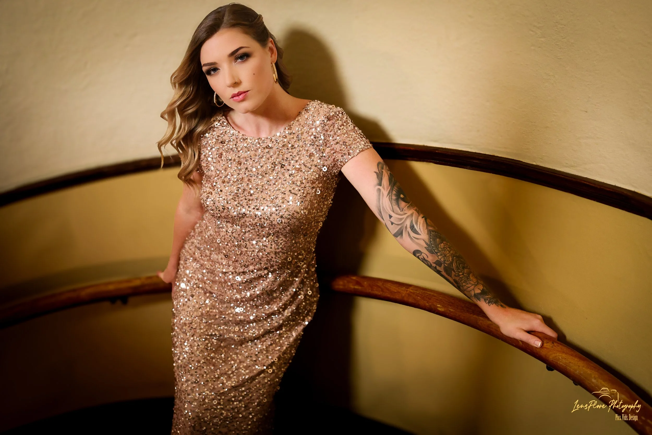 A woman with long wavy hair wearing a sparkling gold dress and earrings, leaning against a curved wooden handrail on a staircase with yellow walls.