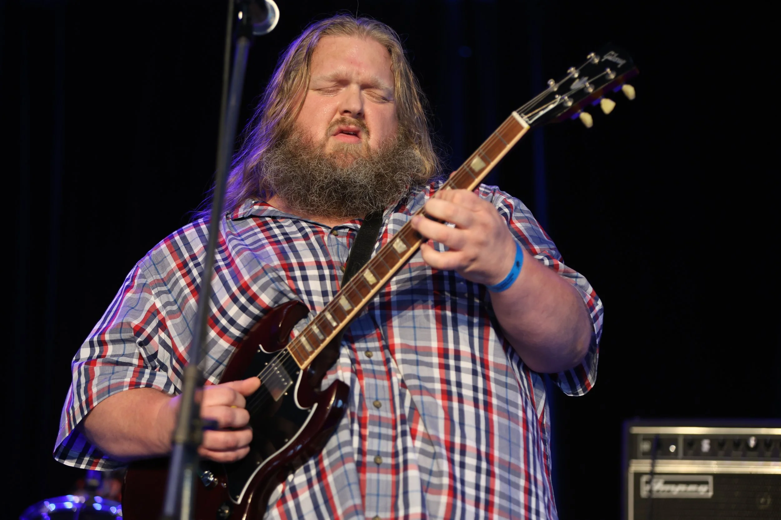 A man with long hair and a beard playing an electric guitar on stage, wearing a plaid shirt.