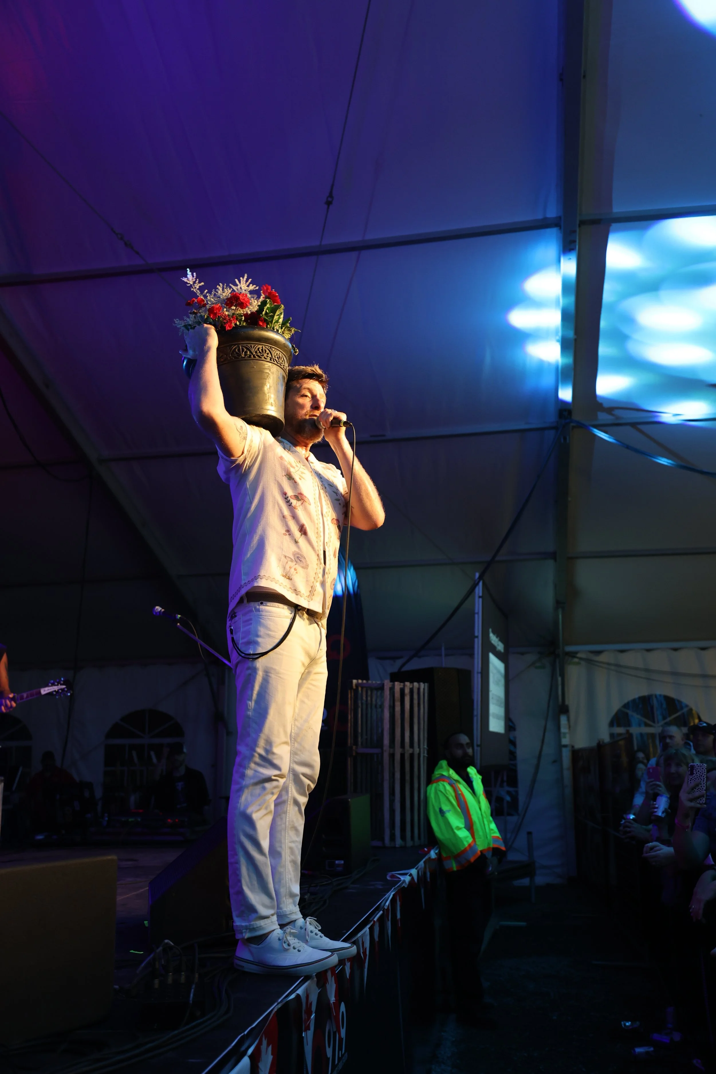 A man on a stage holding a pot of flowers over his shoulder while singing into a microphone, with audience members watching and a security guard nearby.