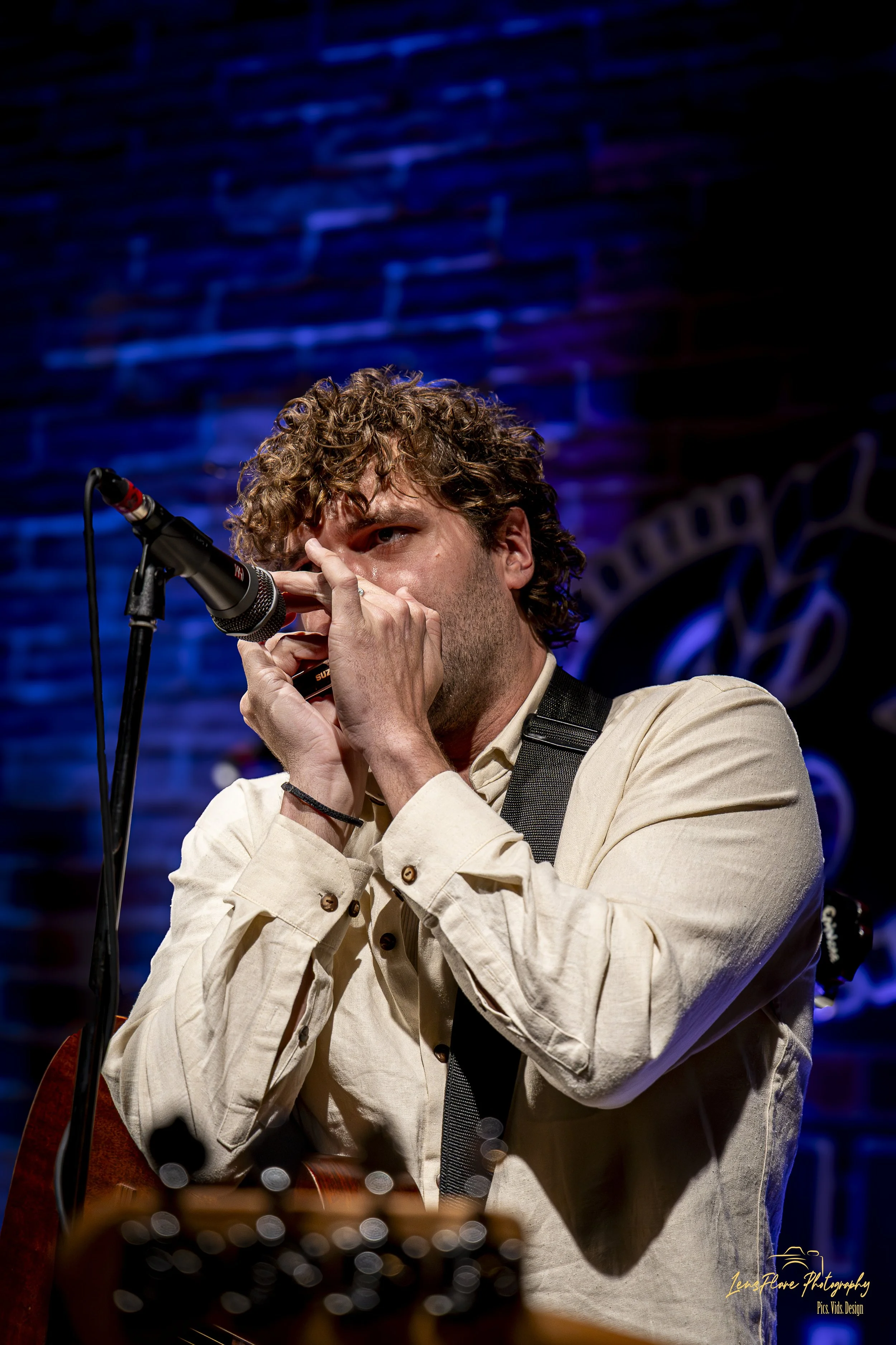 A man with curly hair wearing a light-colored shirt and black suspenders plays a harmonica and sings into a microphone on stage, illuminated by blue and purple stage lighting.