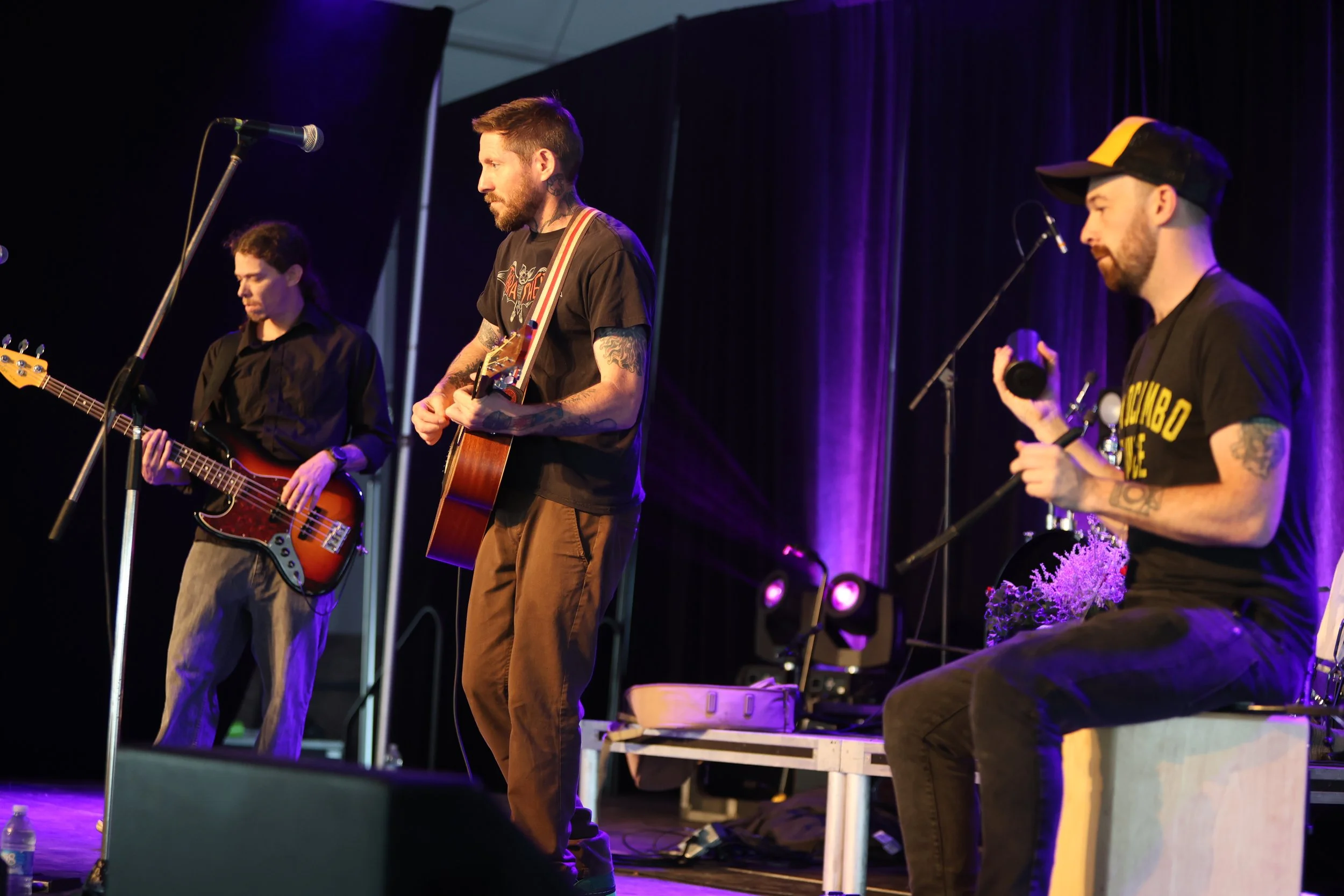 Three musicians performing on stage, with one playing bass guitar, one playing acoustic guitar, and the third sitting with a percussion instrument, all under purple stage lighting.