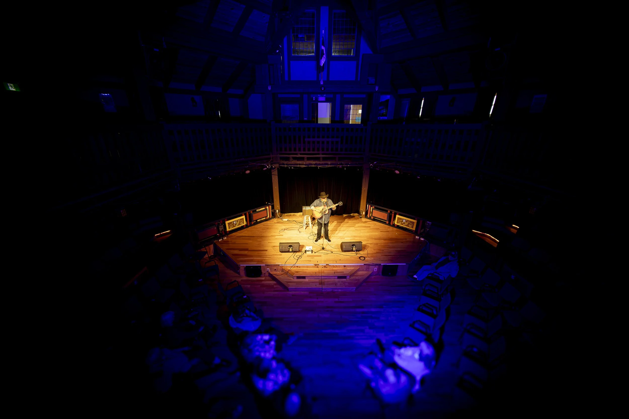 A musician playing an acoustic guitar on stage in a dimly lit auditorium, illuminated by purple and orange lights. The stage is surrounded by an audience watching the performance, with the ceiling and upper balcony visible.