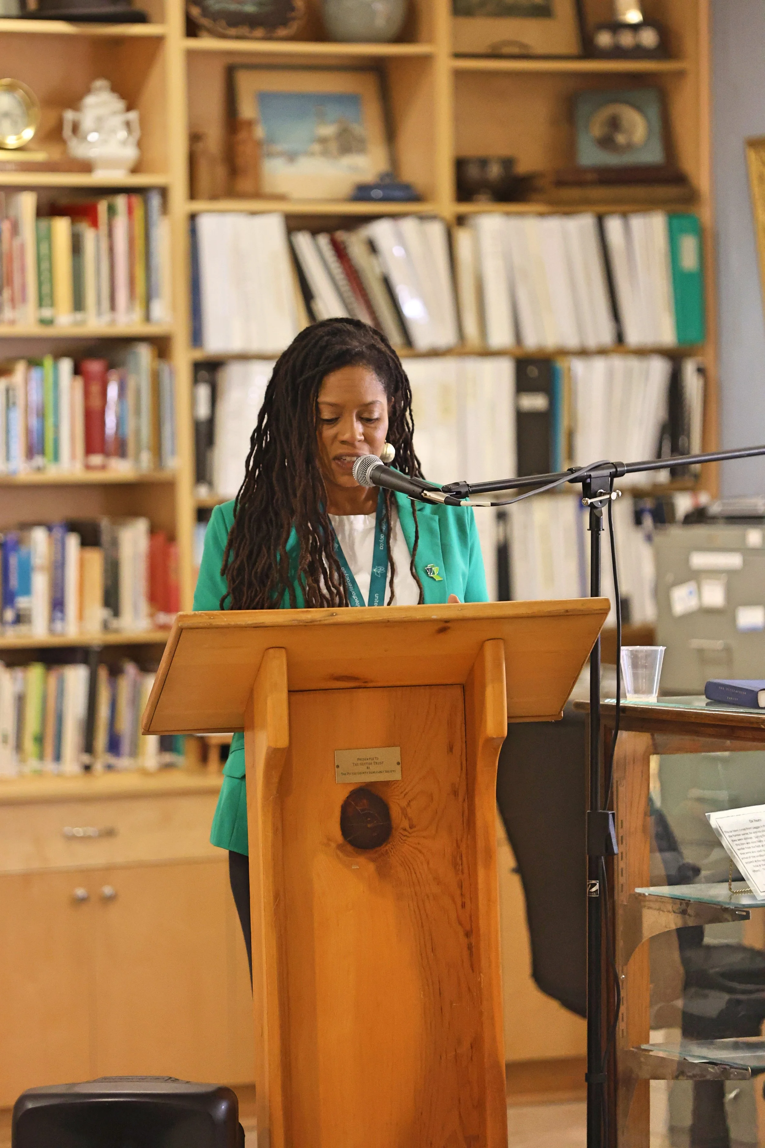 A woman with long dreadlocks is speaking or reading at a wooden podium with a microphone, in a room with bookshelves filled with books and decor.