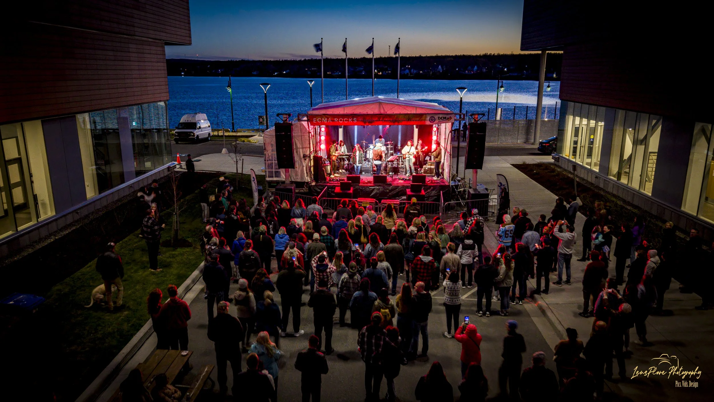 Outdoor concert at sunset by a waterfront with a large audience gathered around a stage and musicians performing under colorful lights.