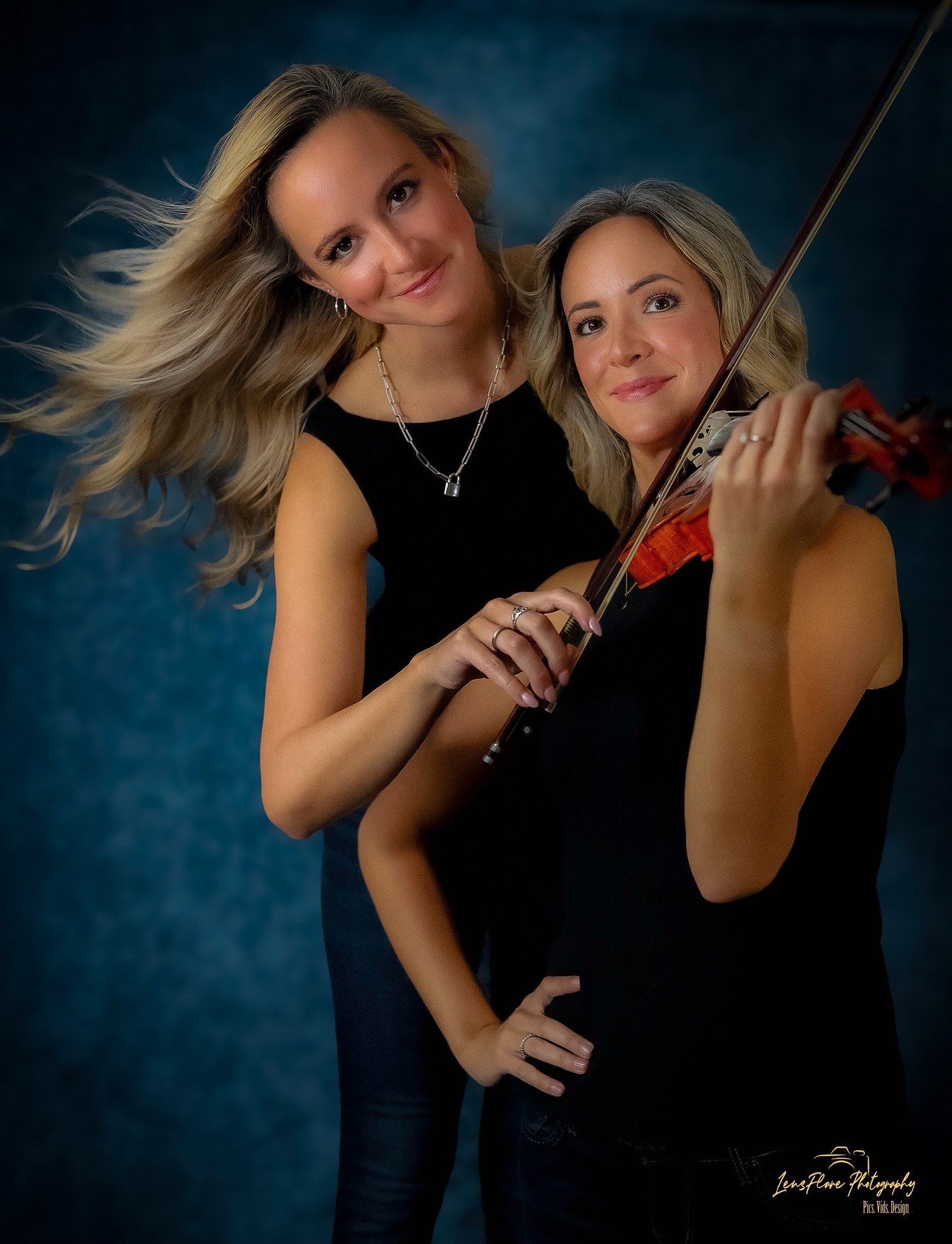 Two women, one with long blonde hair, the other with shoulder-length blonde hair, pose together. The woman with the violin is playing, and the other stands beside her, both smiling at the camera against a dark blue background.