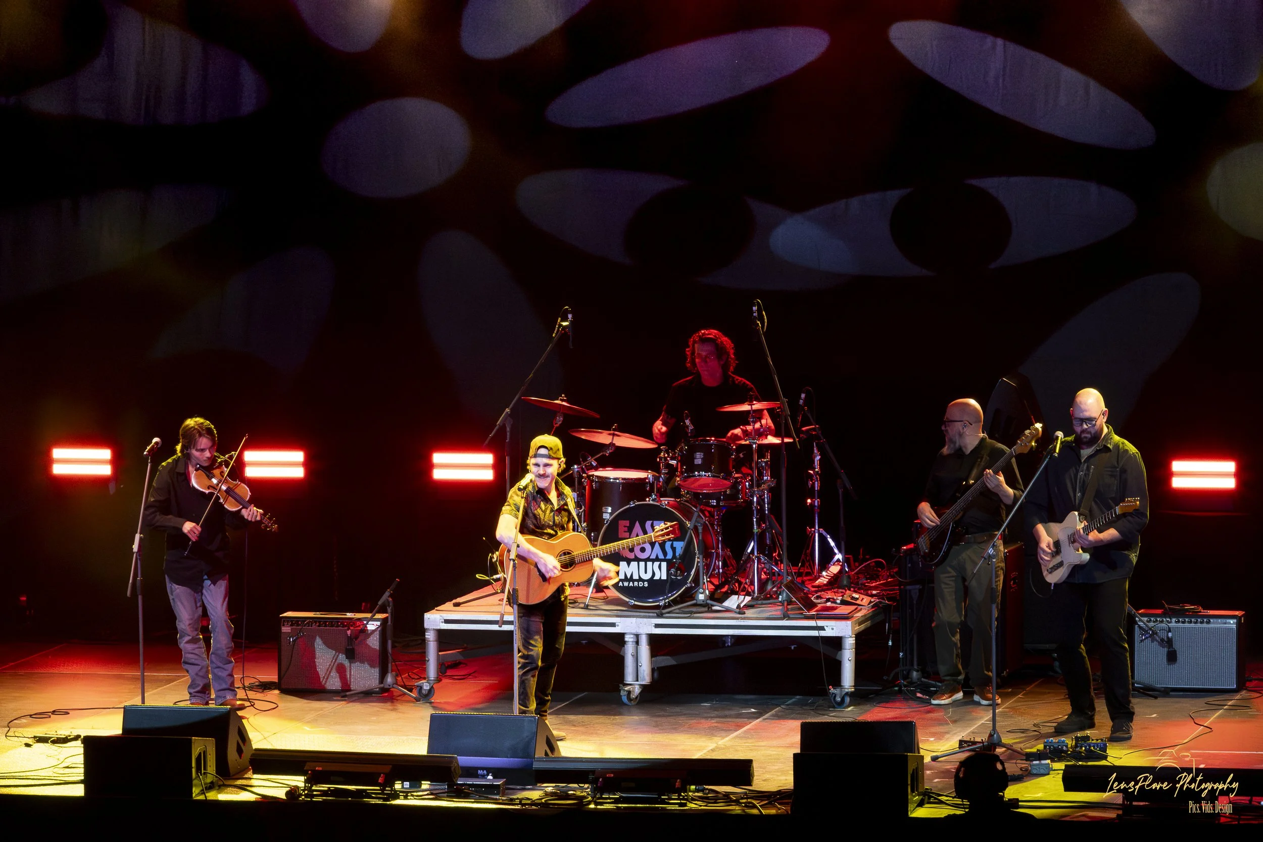 A band performing on stage with a drummer, guitarist, bassist, and violinist. The stage is lit with red lights, and the background features a dark abstract circular pattern. The bass drum has a logo that reads 'East Coast Music Awards.'