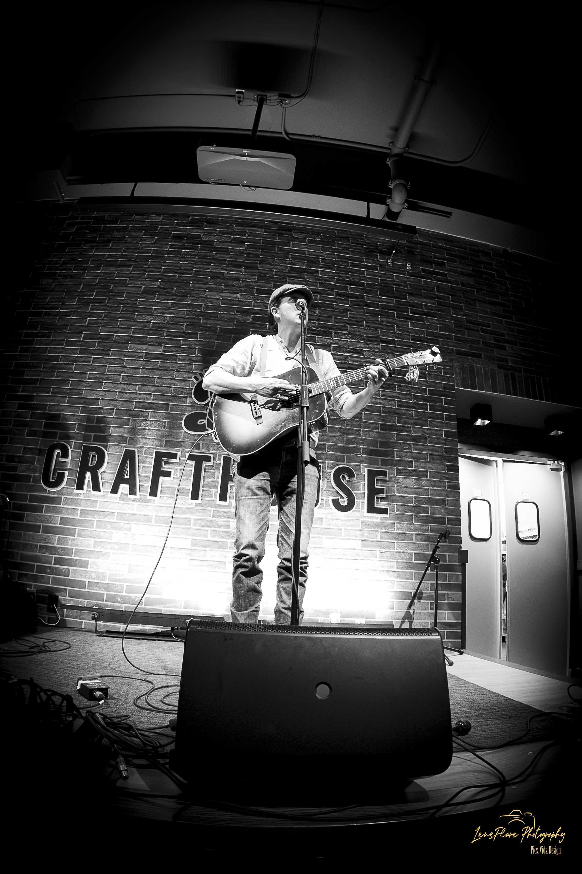 A musician performing on stage with an acoustic guitar in front of a brick wall with the text 'CRAFT' partially visible. The performer is wearing a hat and glasses and is singing into a microphone.