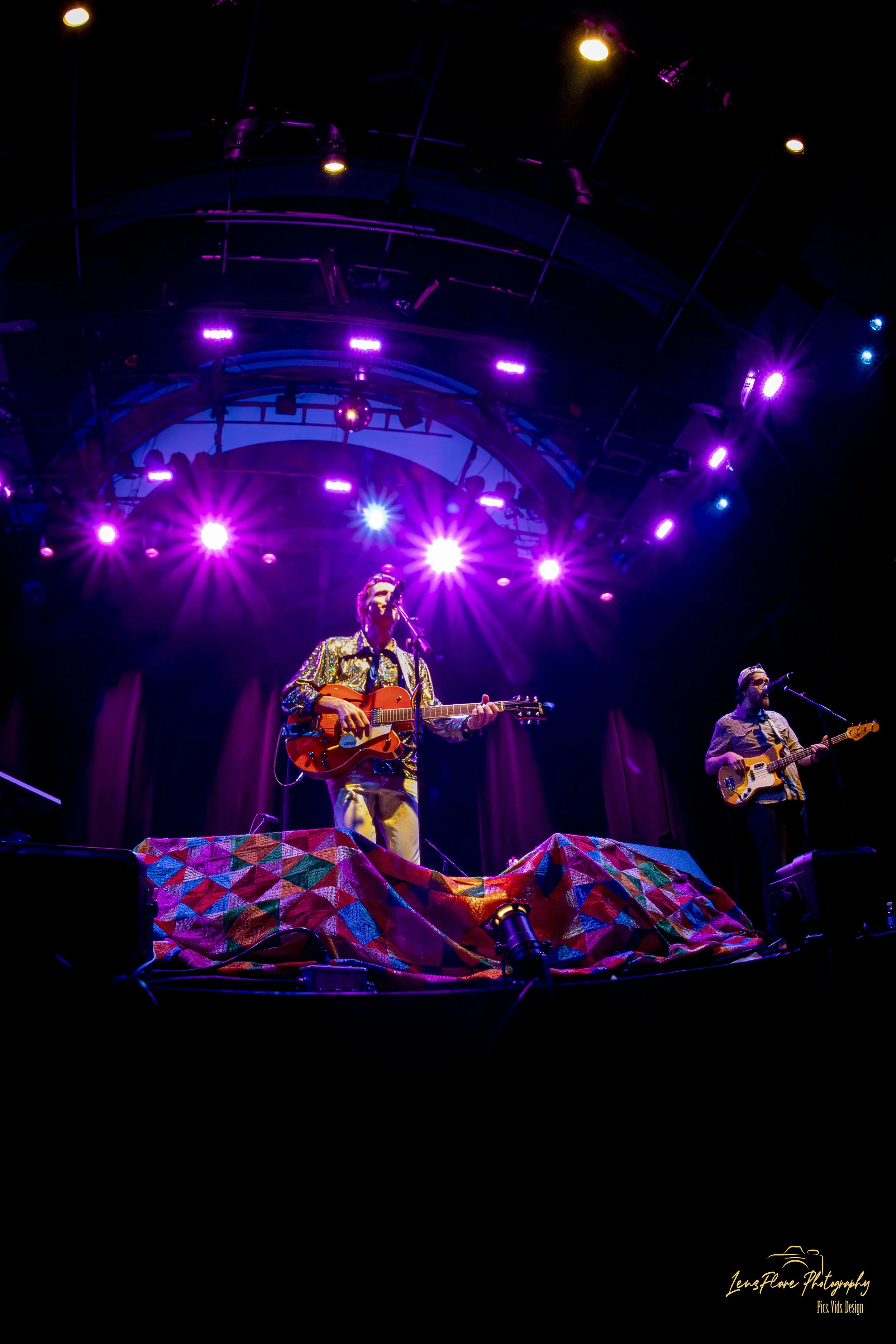 Two musicians perform on a stage illuminated with purple and yellow lights. The musician on the left plays an orange guitar and wears a shiny, patterned shirt. The musician on the right plays a bass guitar, wearing a short-sleeved shirt. The stage ha