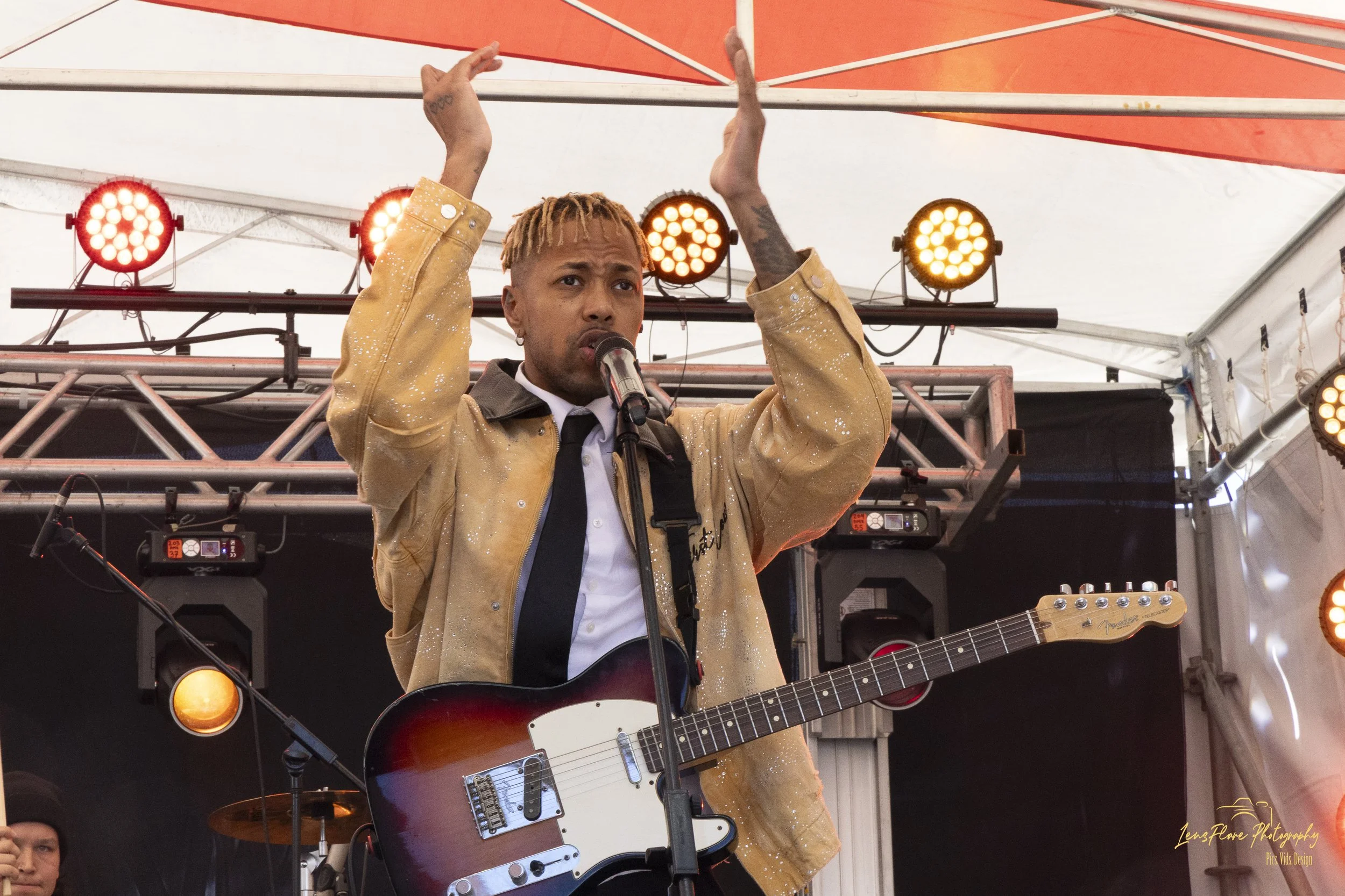 A man with blonde dreadlocks and tattoos is on stage, singing into a microphone, playing an electric guitar, and raising his hands. The stage has orange lights and a white tent canopy overhead.