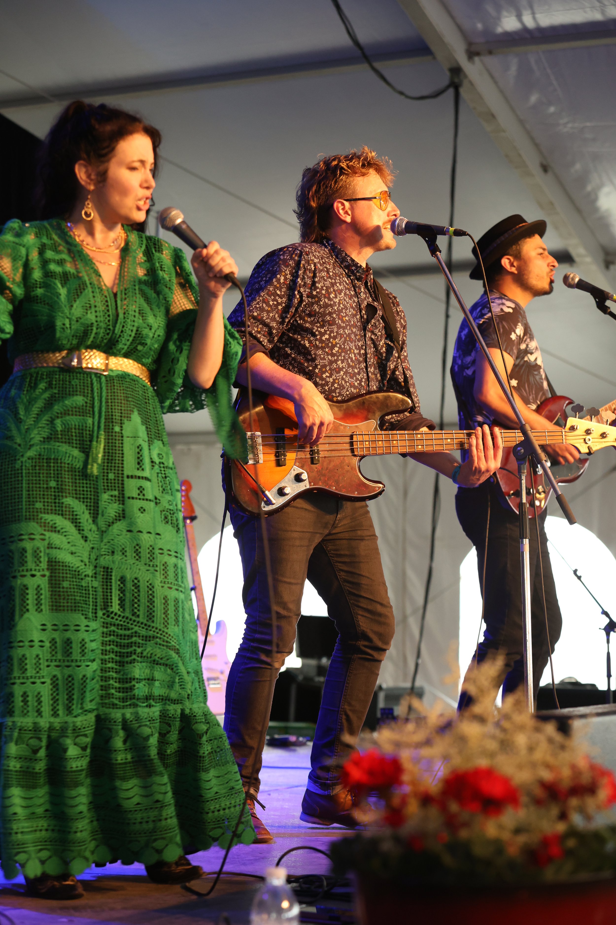 A band performing on stage: a woman in a bright green dress singing into a microphone, a man with glasses and a patterned shirt playing a bass guitar, and another man in a black hat and floral shirt playing an electric guitar.