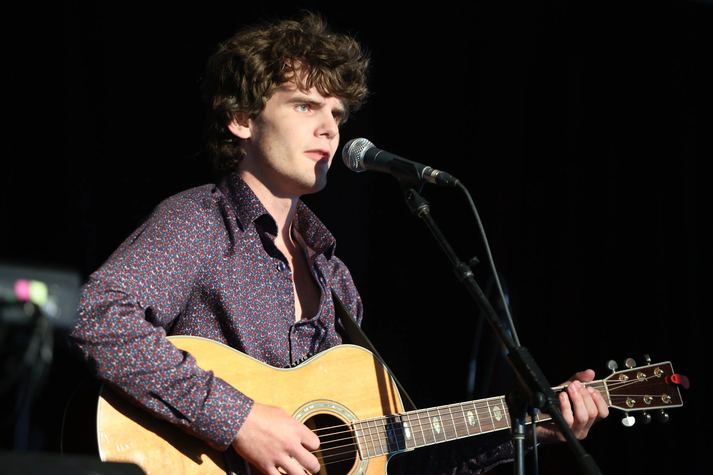 A young man with curly brown hair wearing a patterned shirt is playing an acoustic guitar and singing into a microphone on a dark stage.