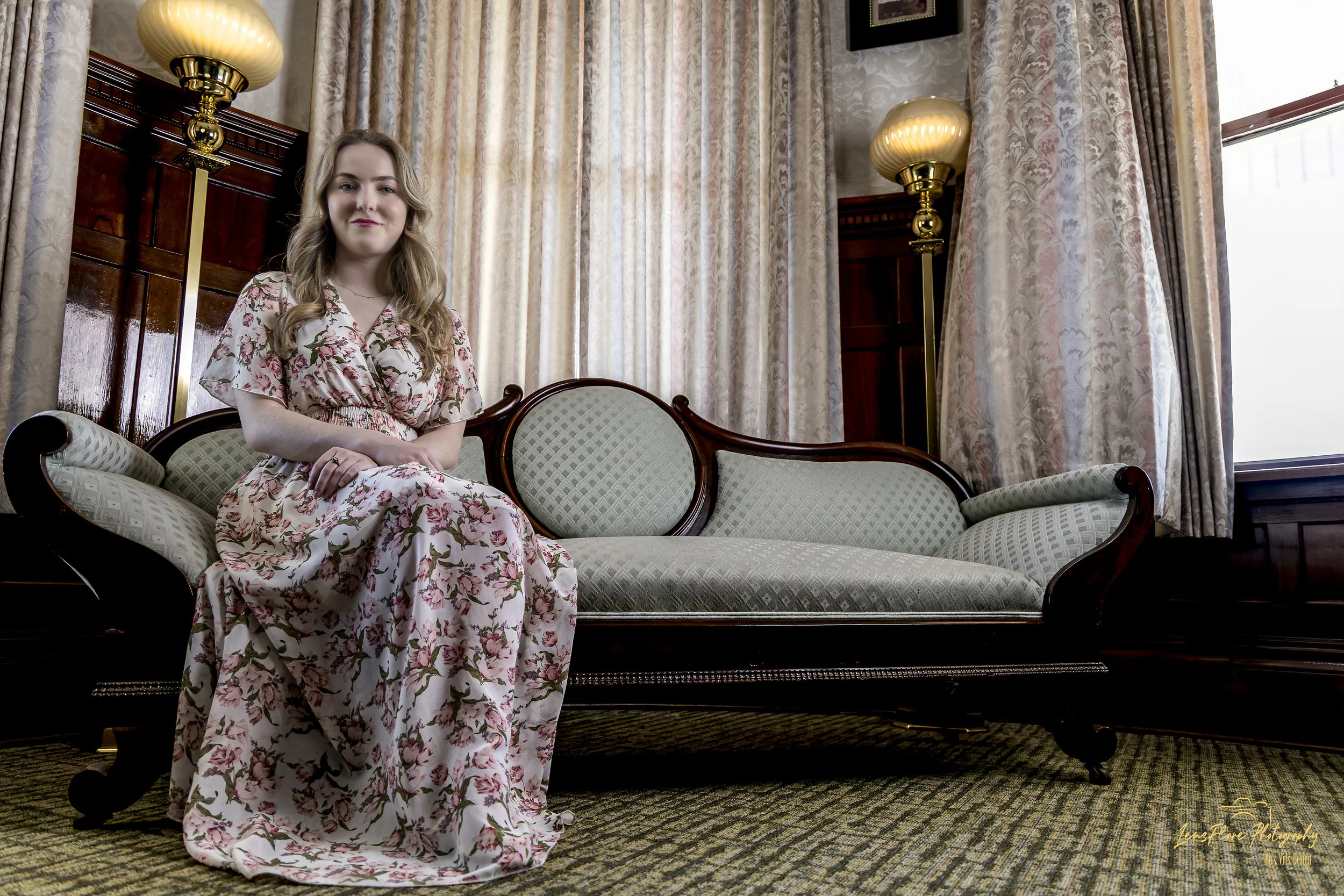A young woman with long blonde hair sitting on a vintage sofa in a room with large patterned curtains, two tall gold-lamp light fixtures, and a large window in the background.