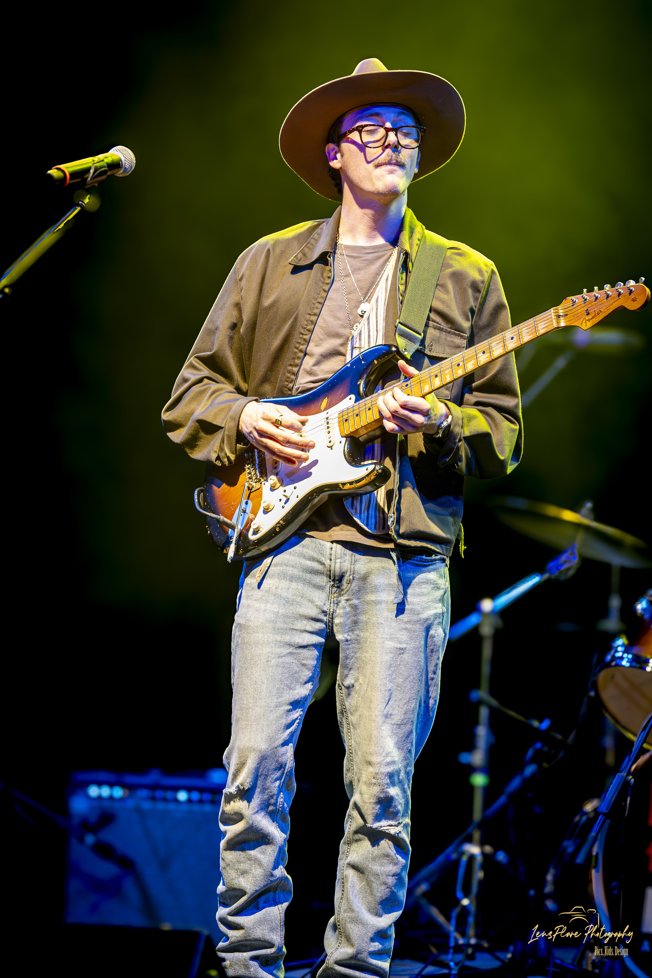 A man wearing glasses, a wide-brimmed hat, a brown jacket, and light-colored jeans is playing an electric guitar on stage with a dark background and stage lighting.