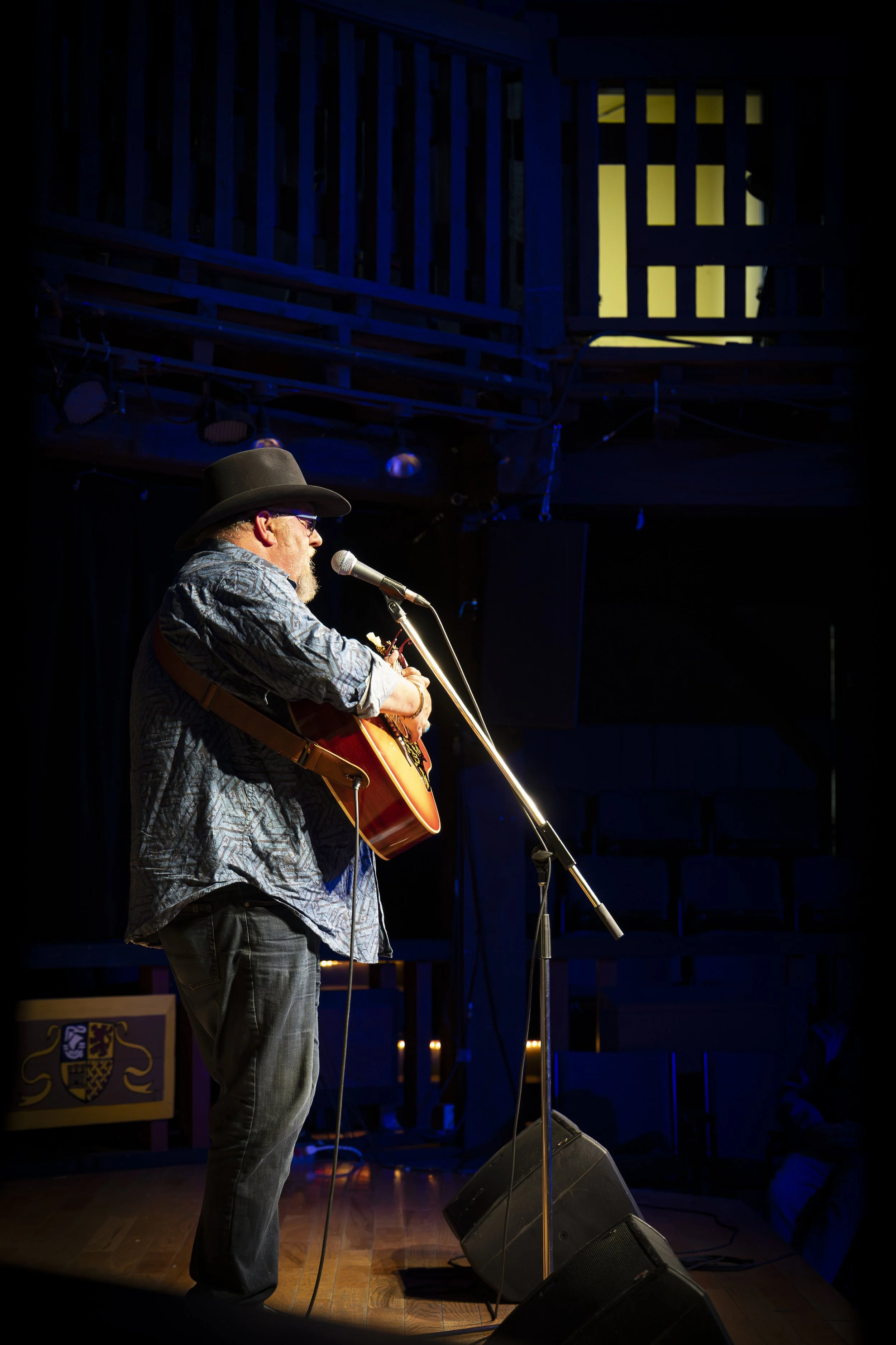 A man with a beard, wearing a cowboy hat, glasses, a patterned shirt, and jeans, playing an acoustic guitar and singing into a microphone on a dimly lit stage with blue and yellow lighting.