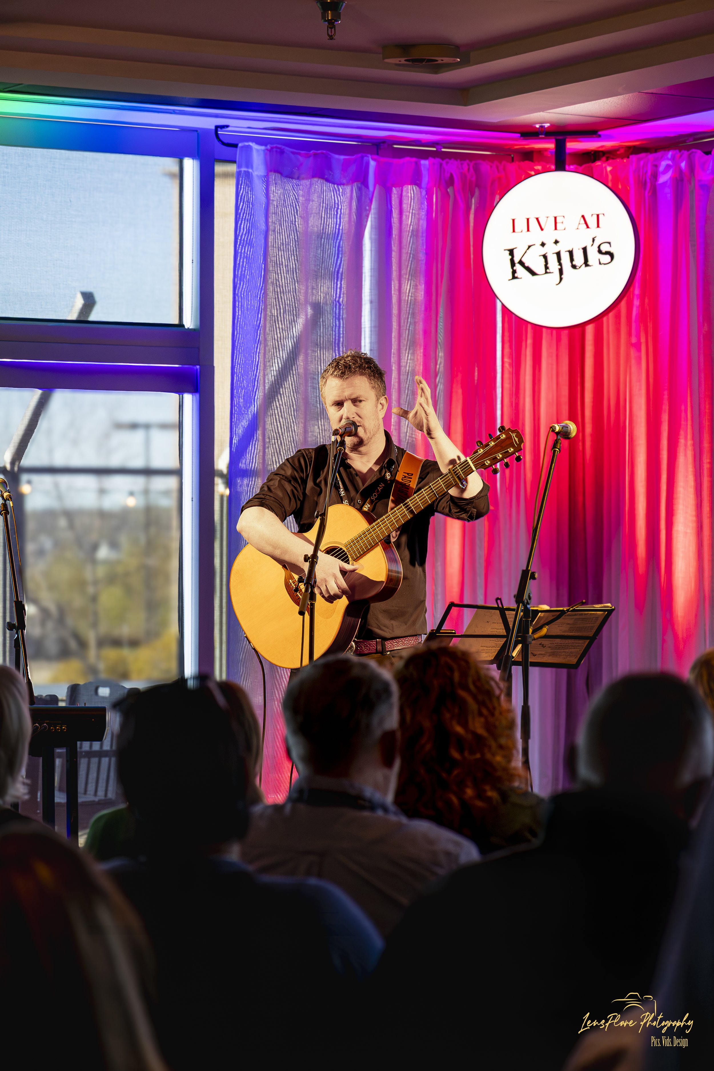 A musician with short hair and a beard is playing an acoustic guitar and singing into a microphone on a stage with pink and purple curtains. A sign above reads 'Live at Kijiu's.' There is an audience visible at the bottom of the image.