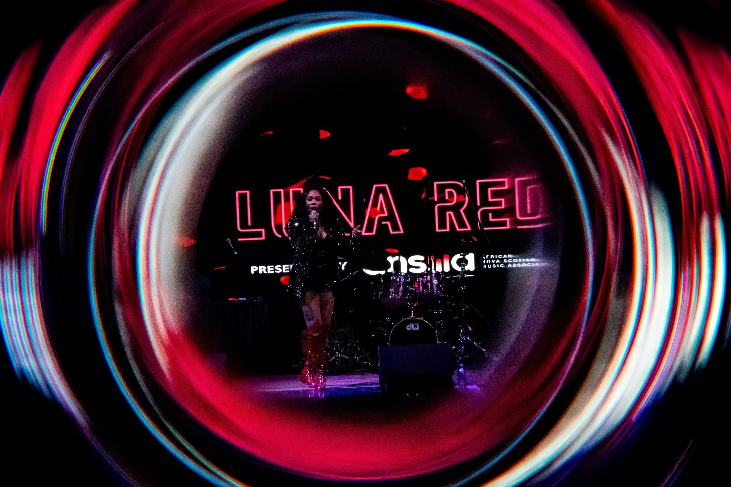 A performer on stage holding a microphone with neon pink letters in the background, framed by colorful swirling lights.