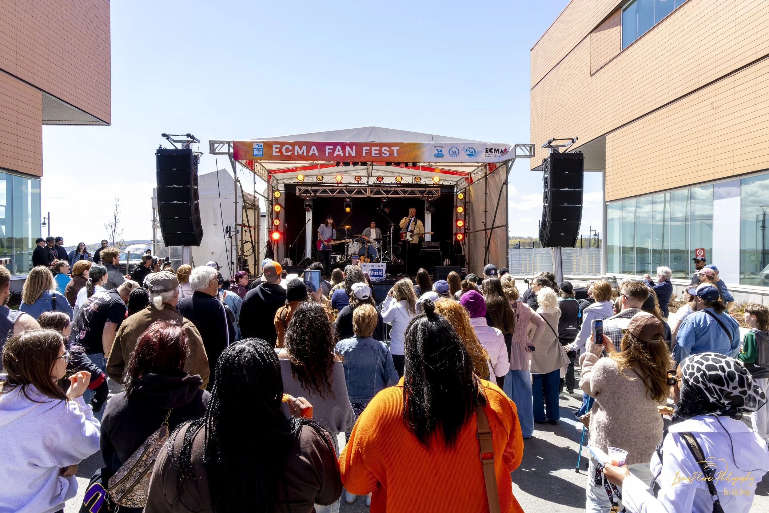 A crowd gathered outdoors watching a band perform on a stage at the ECMA Fan Fest on a sunny day. Modern buildings with large glass windows surround the stage.