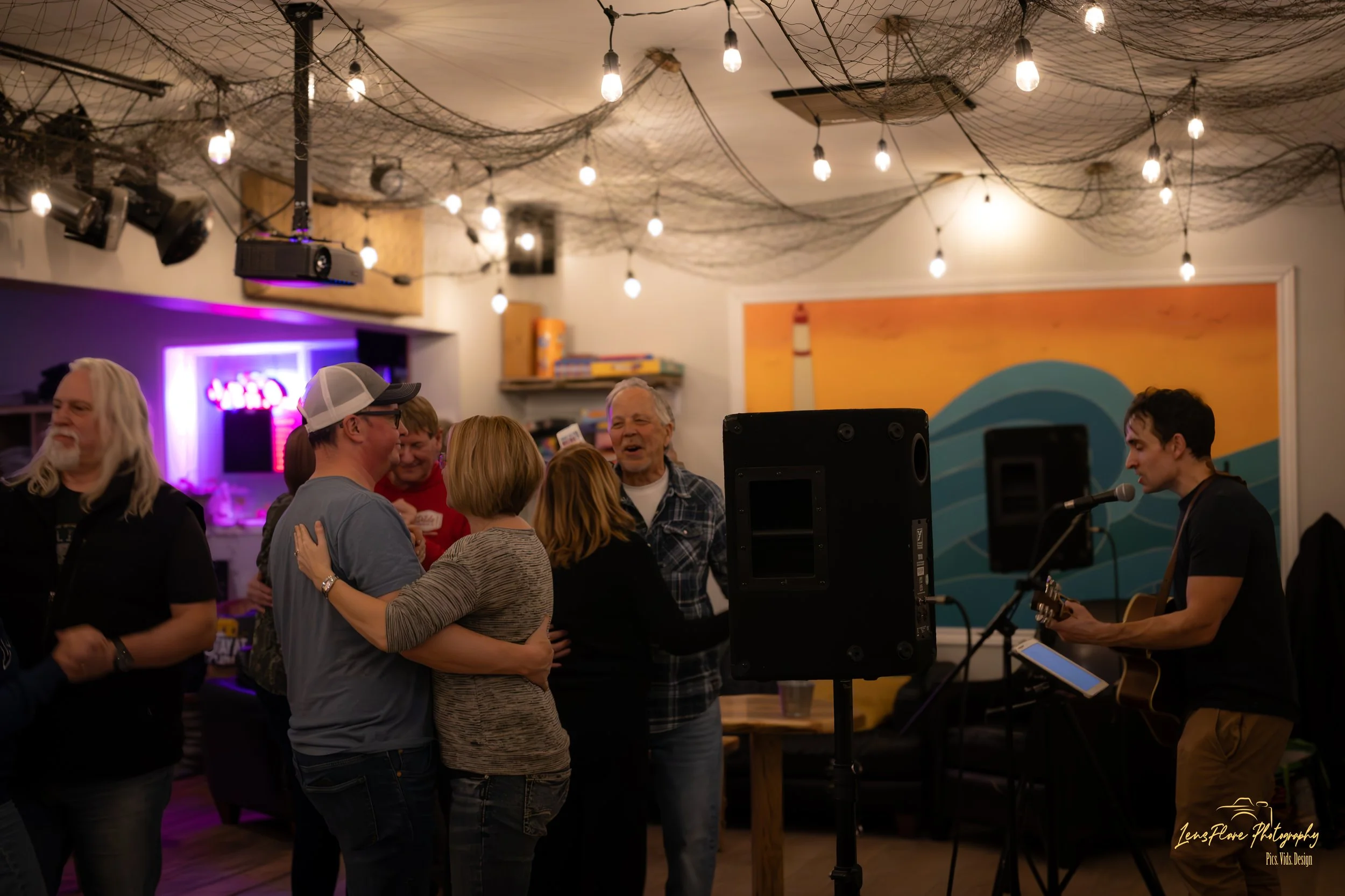 People dancing and singing at a social gathering with a musician playing guitar on stage, under string lights and net decorations, in a cozy indoor setting.