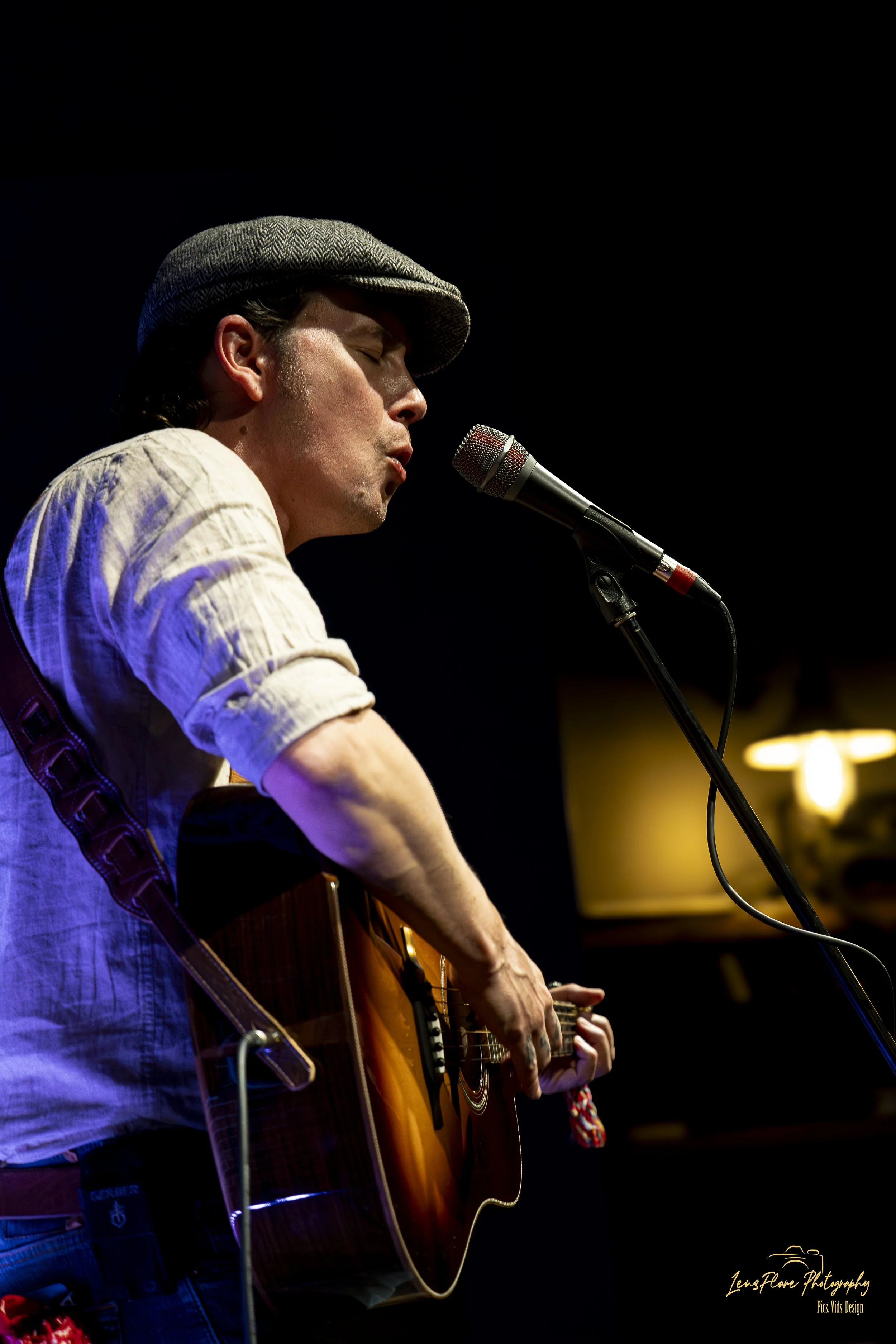 A man wearing a gray hat and beige shirt, singing into a microphone while playing an acoustic guitar on stage with a dark background.