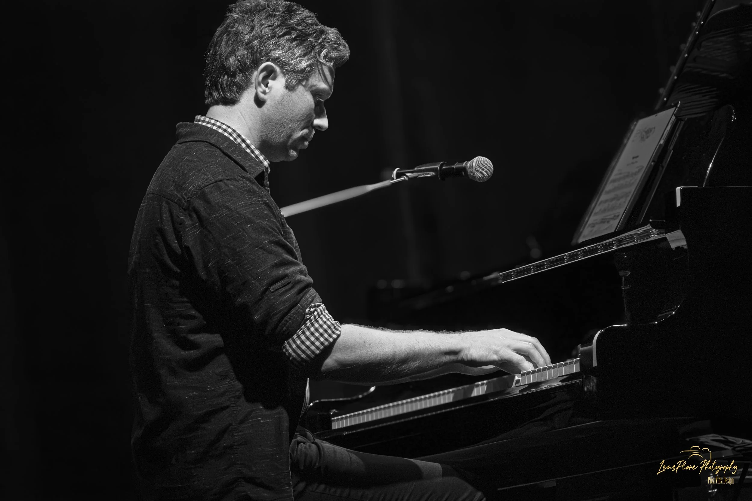 A black and white image of a man with wavy hair playing a grand piano, with a microphone positioned in front of him, illuminated against a dark background.