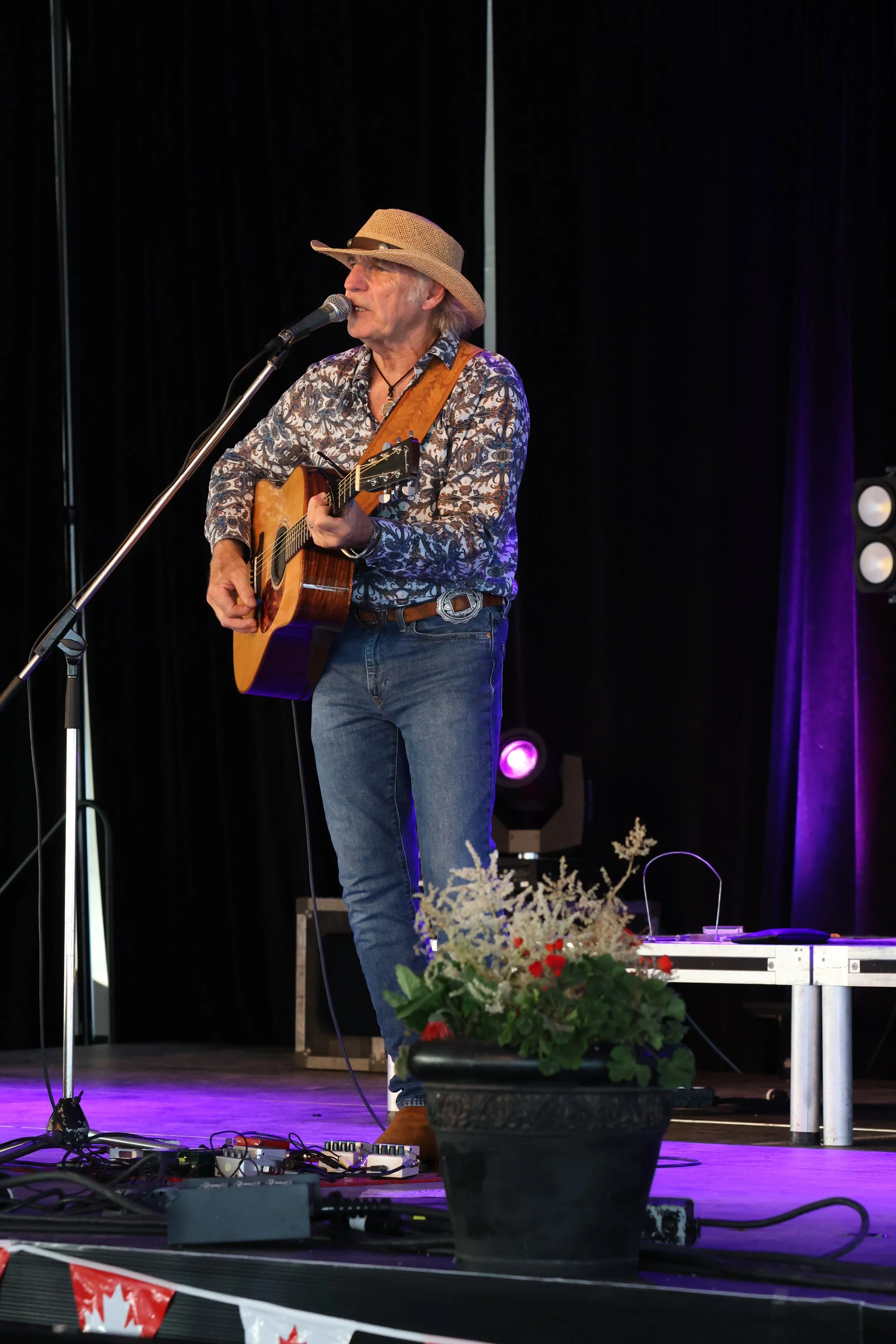 A man wearing a straw hat, patterned shirt, and jeans, playing an acoustic guitar on stage with a microphone while performing.