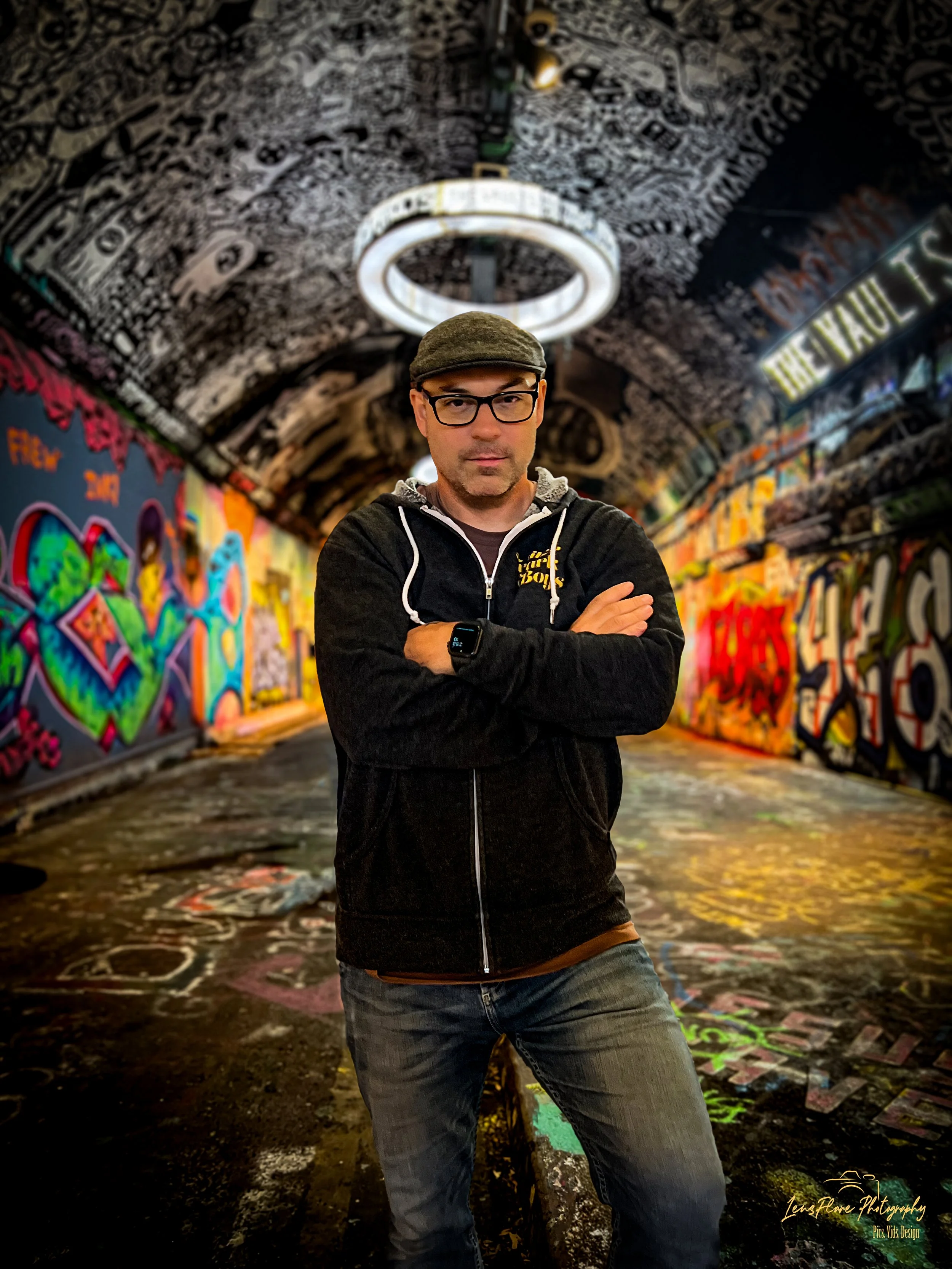 A man standing with arms crossed in a graffiti-covered tunnel with colorful wall art and a circular ceiling light.
