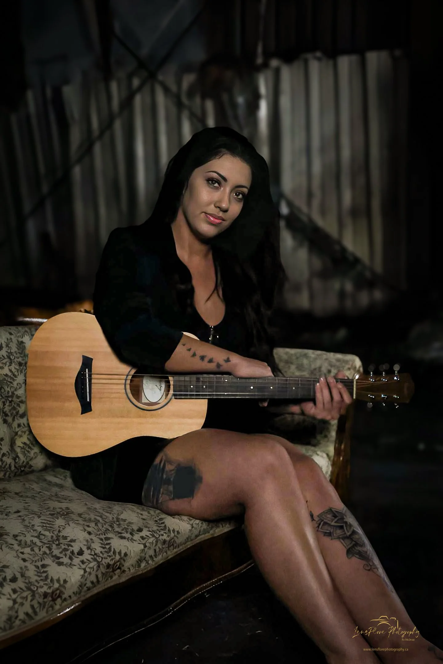 A woman with long dark hair and tattoos on her arm and thigh, sitting on a vintage sofa in a dark, rustic setting, holding an acoustic guitar.