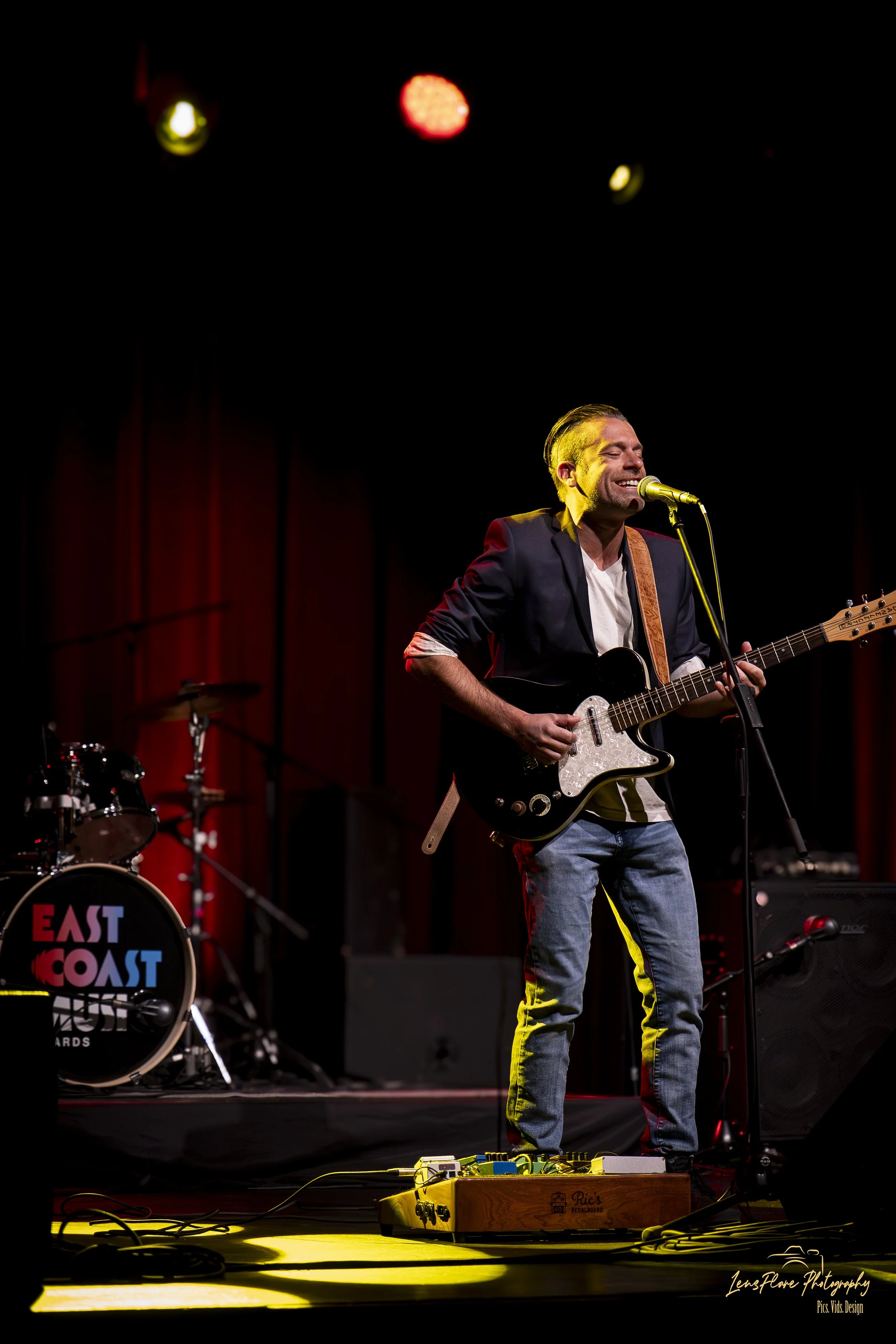 A male musician singing and playing an electric guitar on stage, with a drum set labeled "EAST COAST" in the background, and yellow stage lights illuminating his face and body, while performing in front of black backdrop with stage equipment.