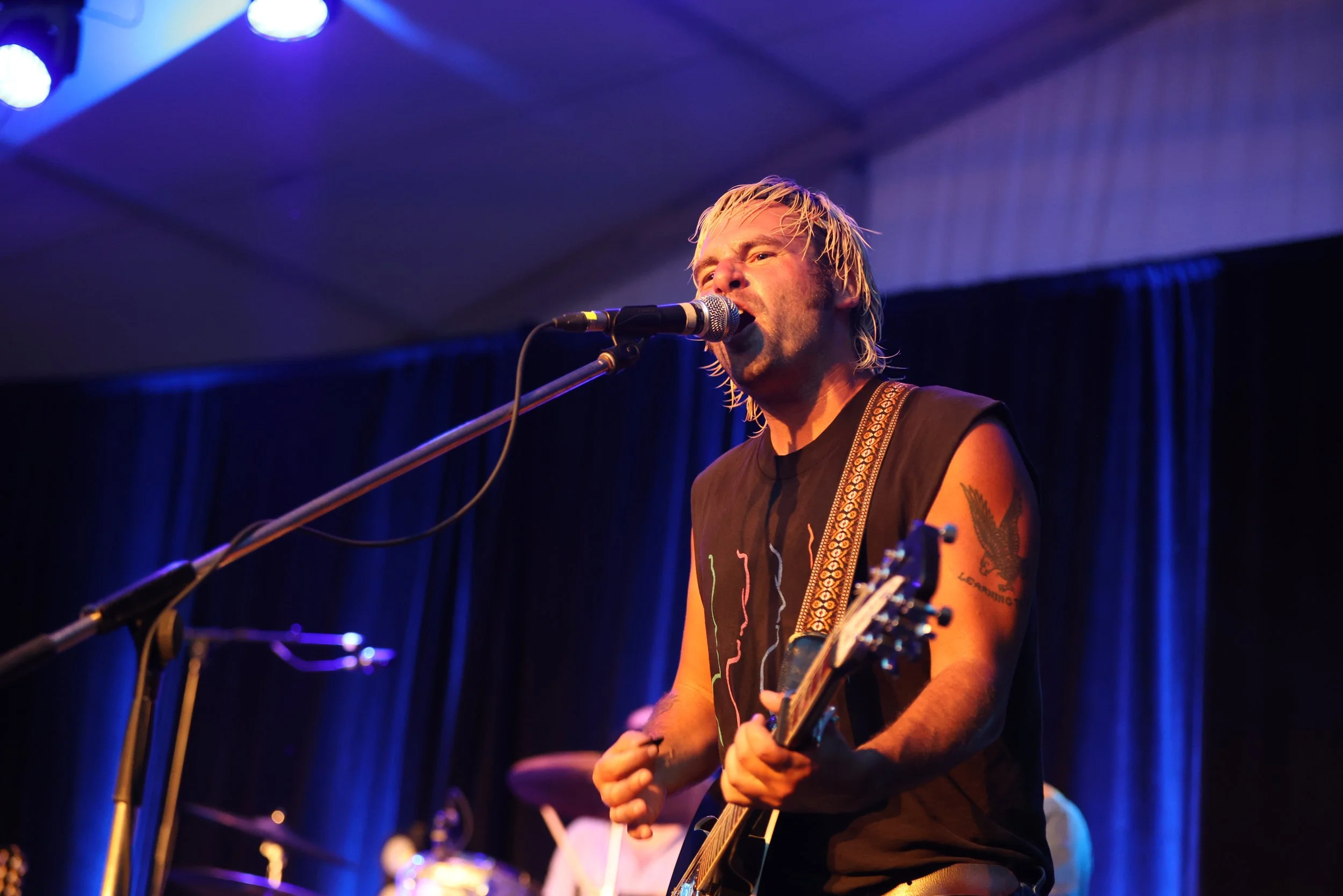 Musician singing into microphone while playing guitar on stage with blue curtains in background.