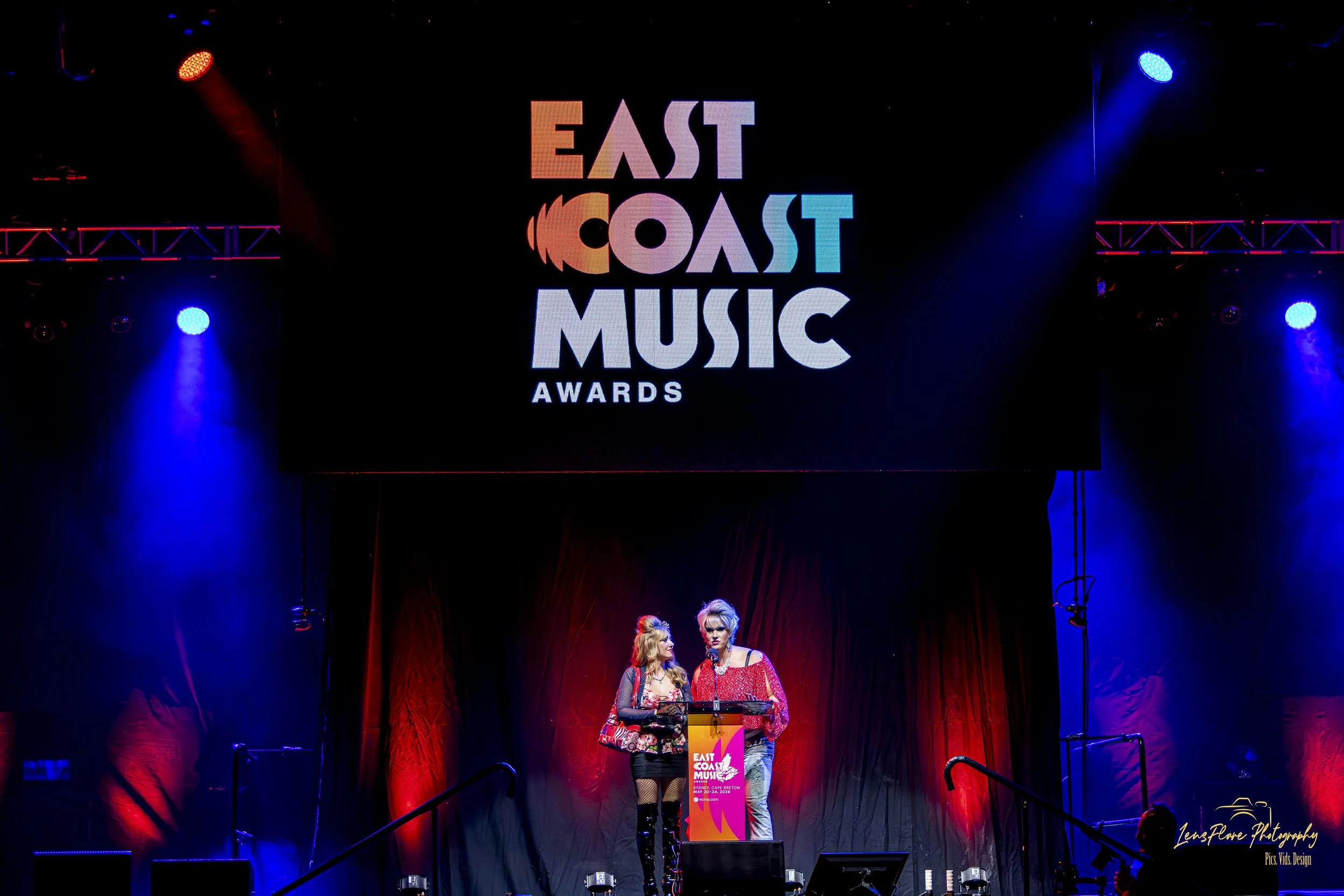 Stage at the East Coast Music Awards with two women speaking at a podium, illuminated by red and blue stage lights.