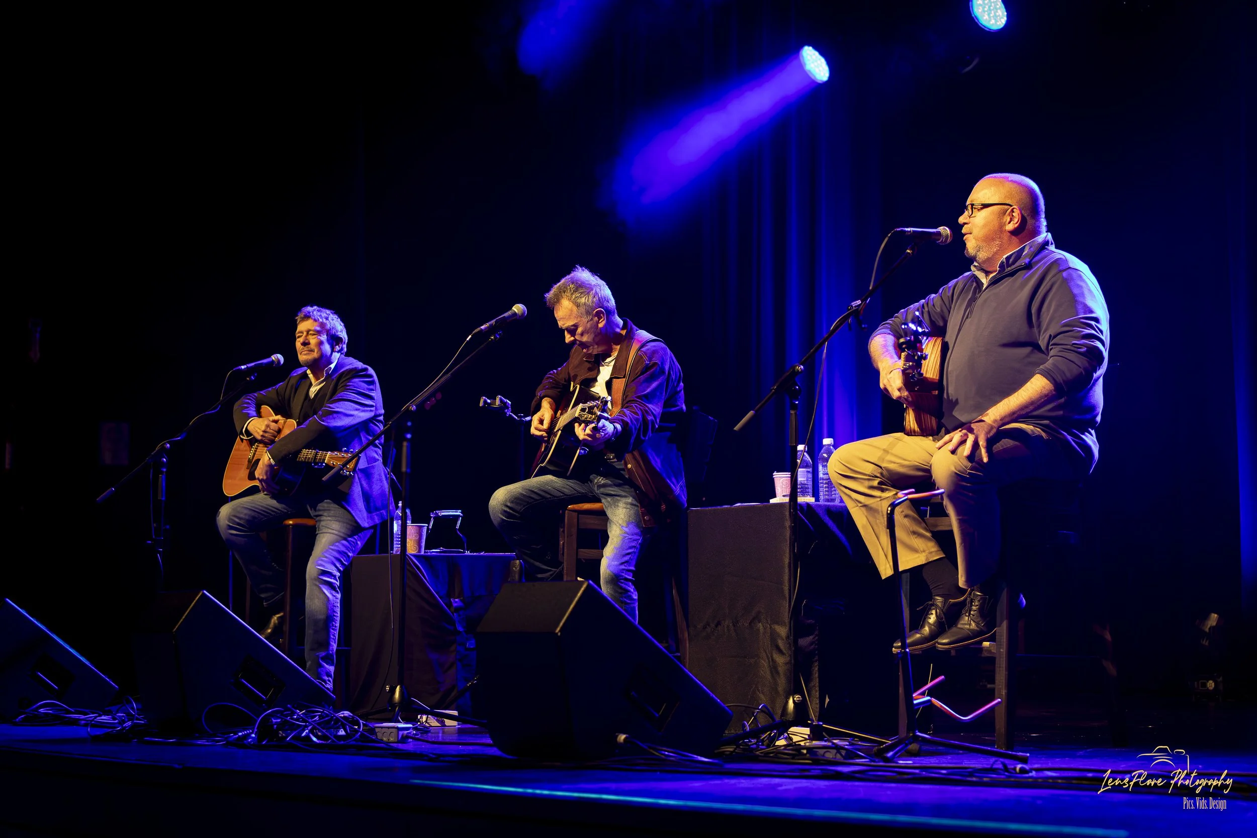 Three male musicians performing on stage with microphones, two playing acoustic guitars, and one holding a microphone. Stage lighting with blue and purple hues.