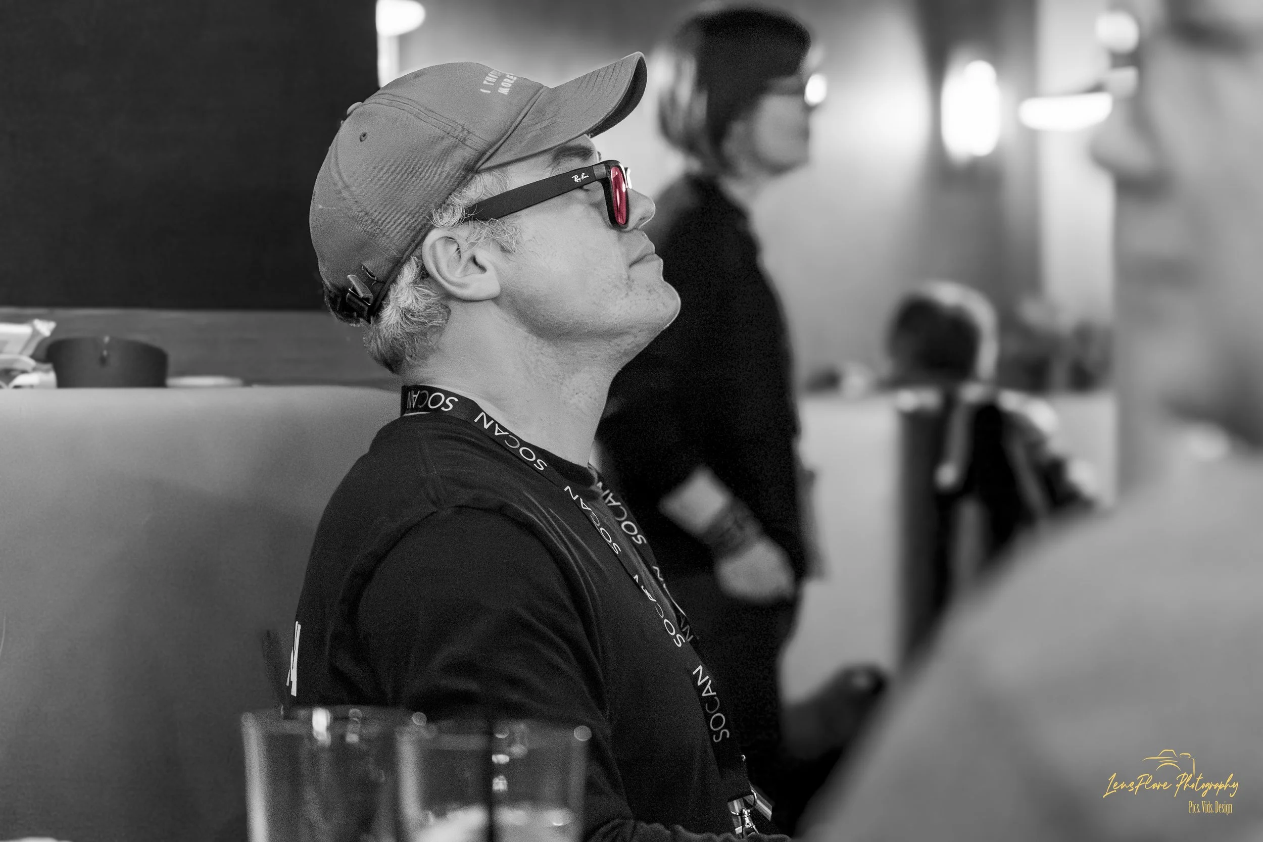 A person with curly hair wearing sunglasses, a baseball cap, and a lanyard with the word 'SOCIAL' on it, sitting on a bench or chair indoors. They appear to be relaxing or observing something. The scene is in black and white, with blurred figures in 