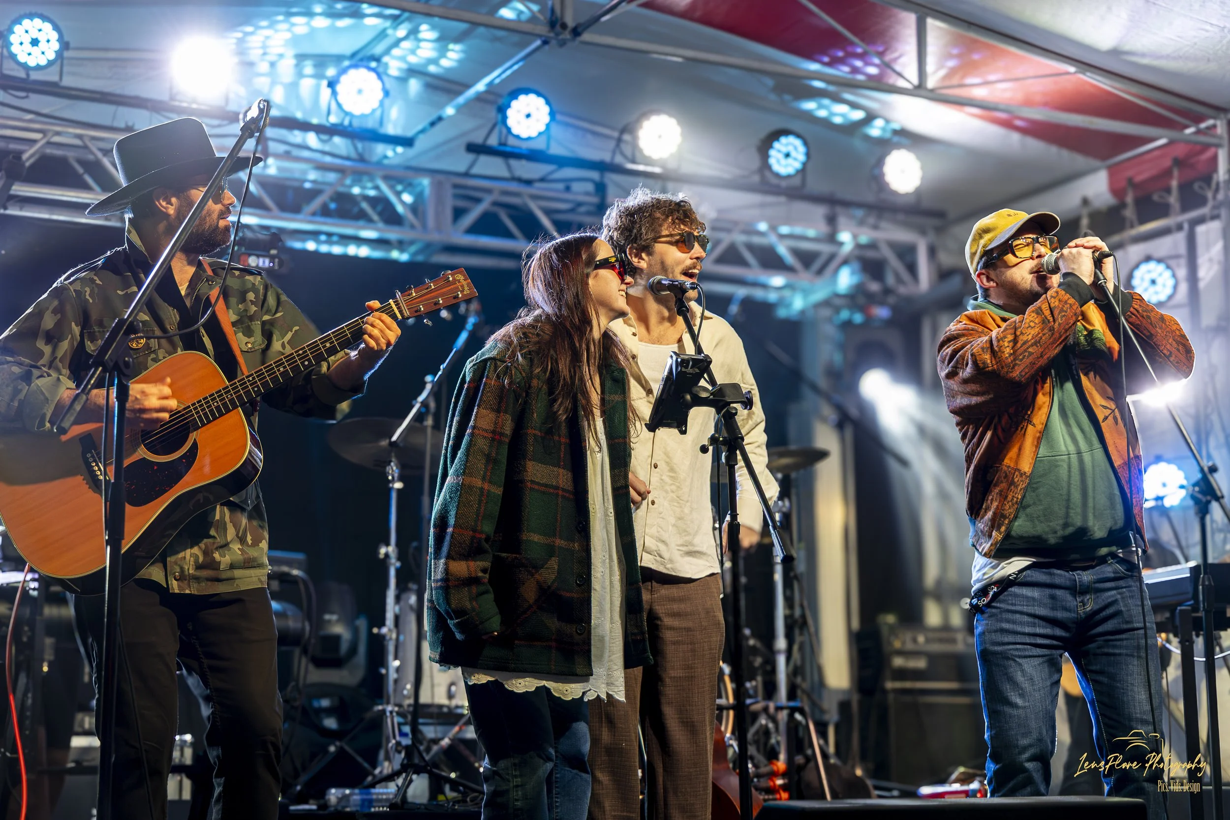 A group of musicians performing on stage at a concert; they include a guitarist wearing a camouflage jacket and hat, and a singer wearing sunglasses and colorful clothing, with stage lighting above.