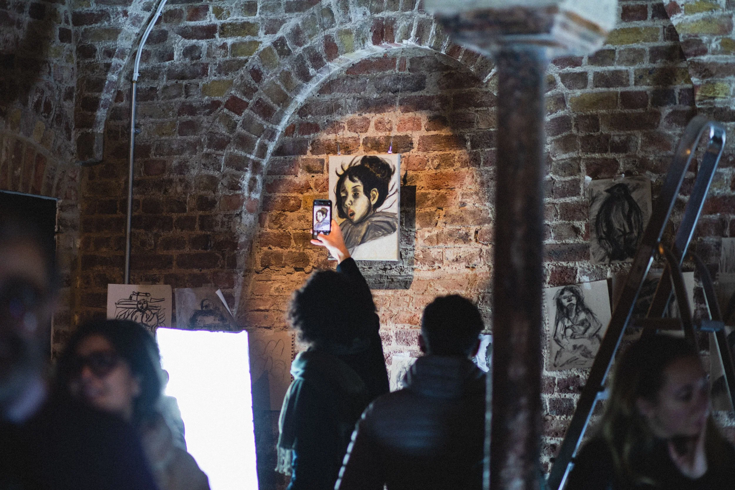Open studio of Malak Mattar. People viewing and taking photos of artwork displayed on a brick wall inside an art gallery or studio, with sketches and paintings surrounding them.