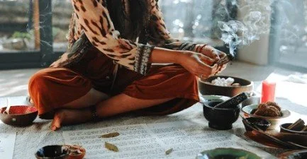 A person sitting cross-legged on a floor with incense and various bowls of food or offerings around them, near a window.