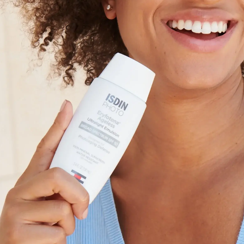A woman with curly hair smiling while holding a white bottle of ISDIN photo sunscreen close to her face.
